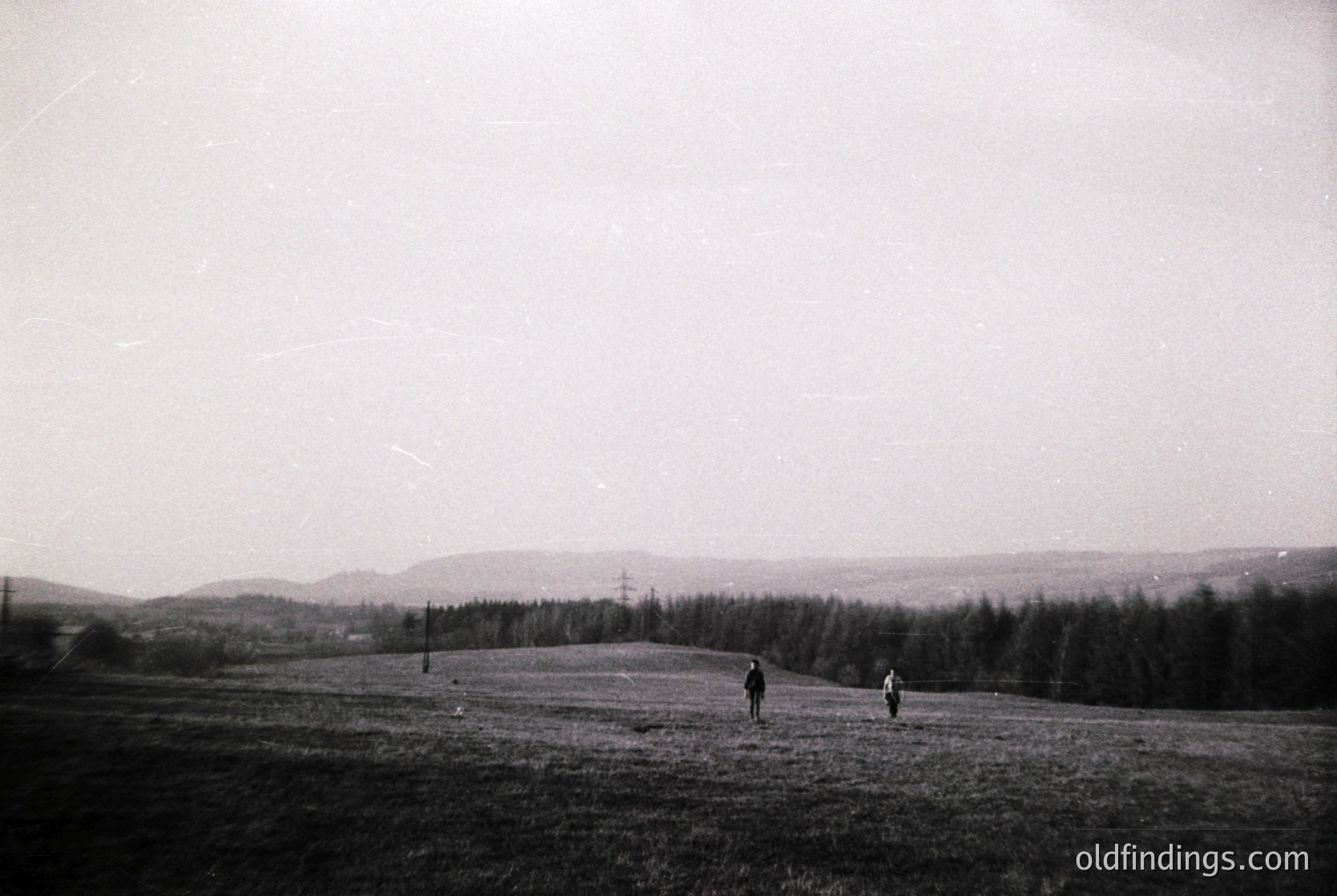Black-and-white landscape featuring two figures walking across an open, slightly hilly field. Dense forest lines the horizon, with distant power lines and a faint industrial structure visible. Mid-20th century rural or agricultural setting.