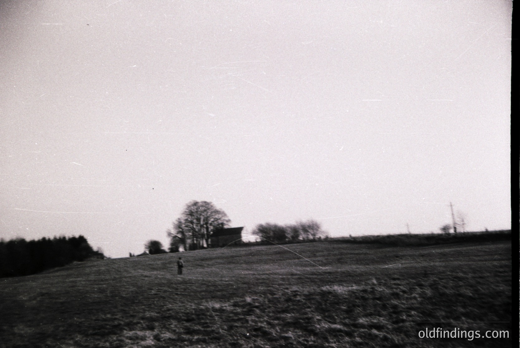 Vintage black-and-white rural landscape featuring a lone figure in mid-stride across an open field. Distant farmhouse and leafless trees suggest late autumn/winter. Horizontal composition highlights expansive horizon. Ideal for historical or nostalgic stock imagery.
