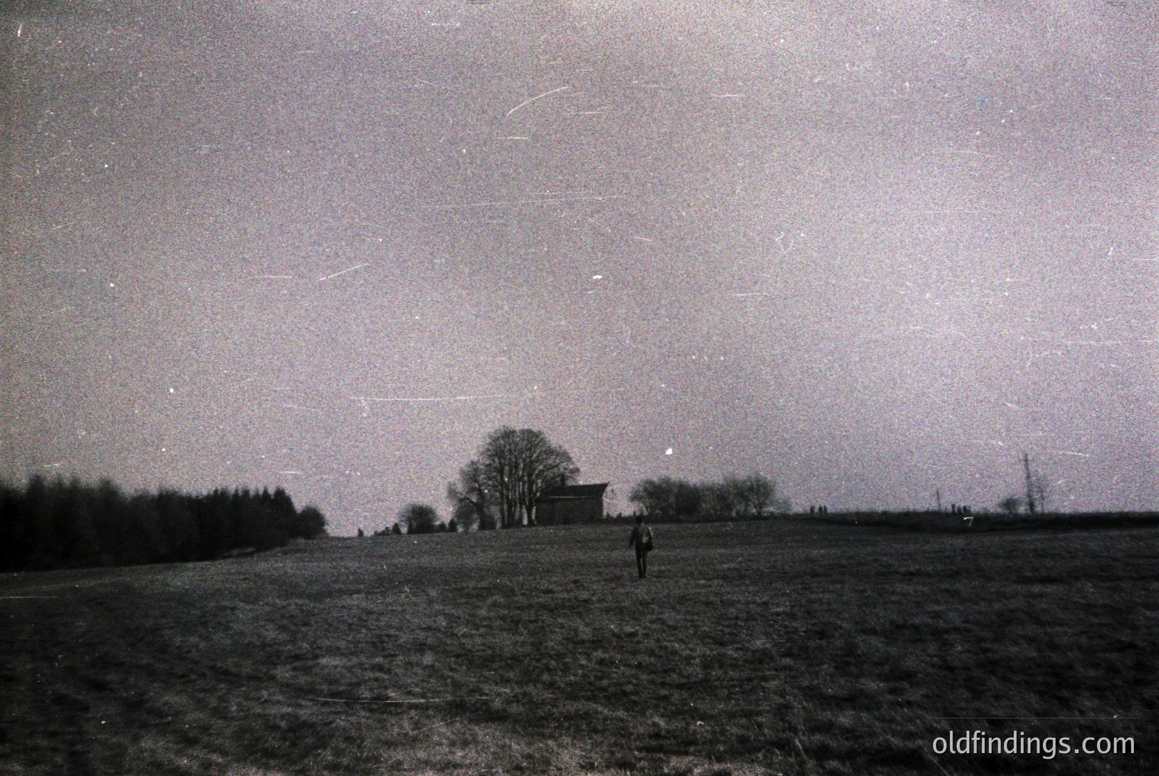 Black-and-white nighttime landscape featuring a lone figure standing in an open field under a starry sky with visible meteor trails. A solitary tree and small building frame the scene, suggesting rural isolation. Likely mid-20th century due to photographic style and attire.