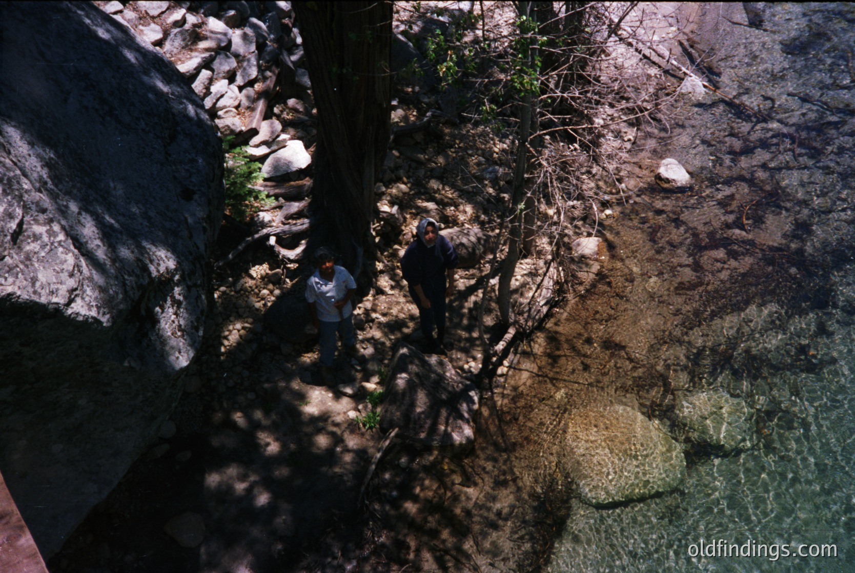 Aerial view of two hikers navigating a shallow, rocky riverbed surrounded by dense forest. Sunlight filters through tree branches, casting dappled shadows. Likely a temperate climate, possibly North America or Europe.