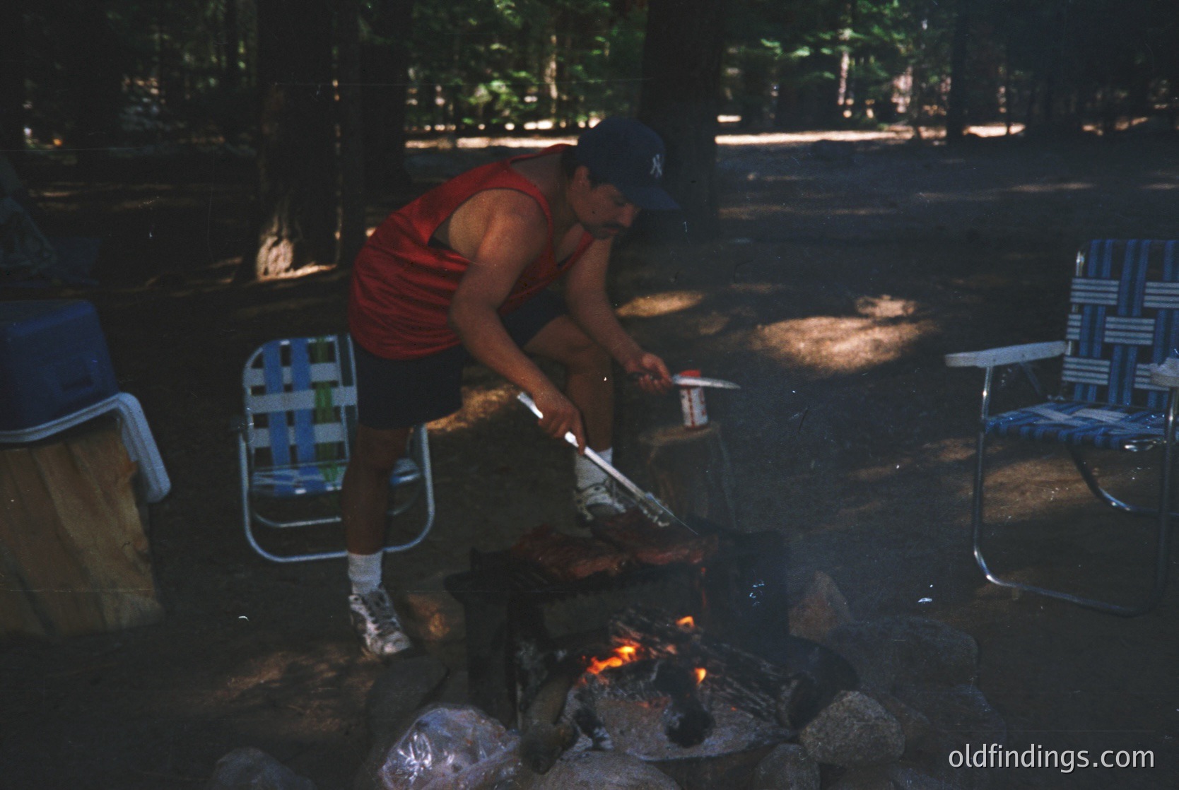 Man grills skewered meat over an open fire in a shaded outdoor picnic area. Foldable metal chairs and a cooler sit nearby. Warm, golden lighting suggests late afternoon.