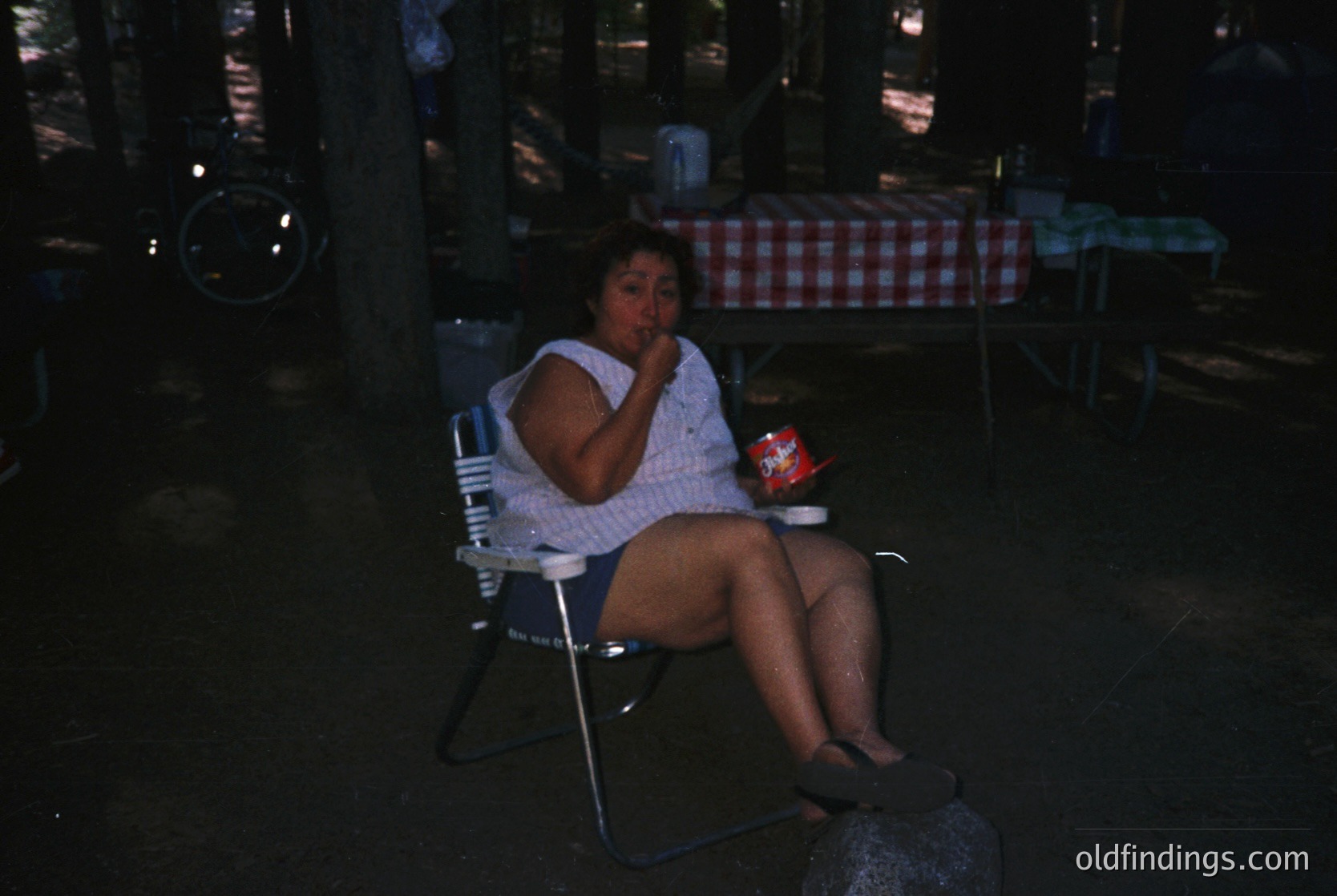 Vintage black-and-white photo of a woman seated on a metal chair in a dimly lit outdoor café, holding a small red container. Checkered red-and-white tablecloth and industrial-style lighting fixtures visible. Likely mid-20th century, urban setting. éCulture