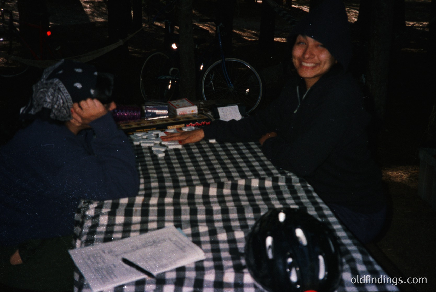 Vintage diner scene with two patrons seated at a checkered table, illuminated by warm overhead lights. One wears a hooded jacket, the other a knit cap. A menu, salt/pepper shakers, and a coffee cup sit on the table. Likely 1970s–1980s American diner setting.