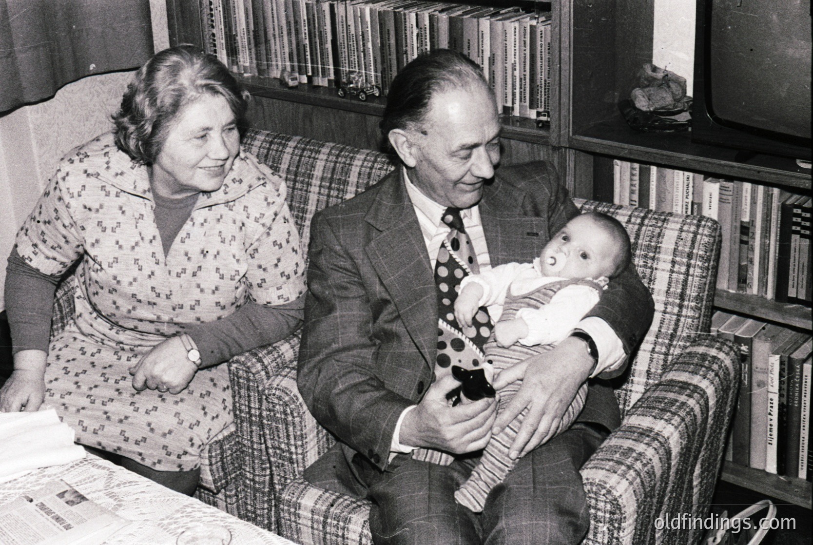 Mid-20th century (1950s-1960s) black-and-white family portrait: elderly couple in formal attire—man in suit & tie, woman in floral dress—holding a baby in a plaid armchair. Bookshelves and vintage decor suggest a middle-class home. Warm, candid moment captures generational bonds.
