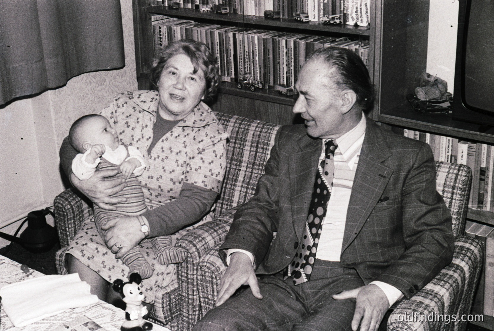 Mid-20th century (likely 1950s–1960s) indoor portrait: elderly couple and infant in a cozy living room with floral-patterned armchairs, a bookshelf filled with bound volumes, and a teddy bear. Formal attire suggests a posed family moment.