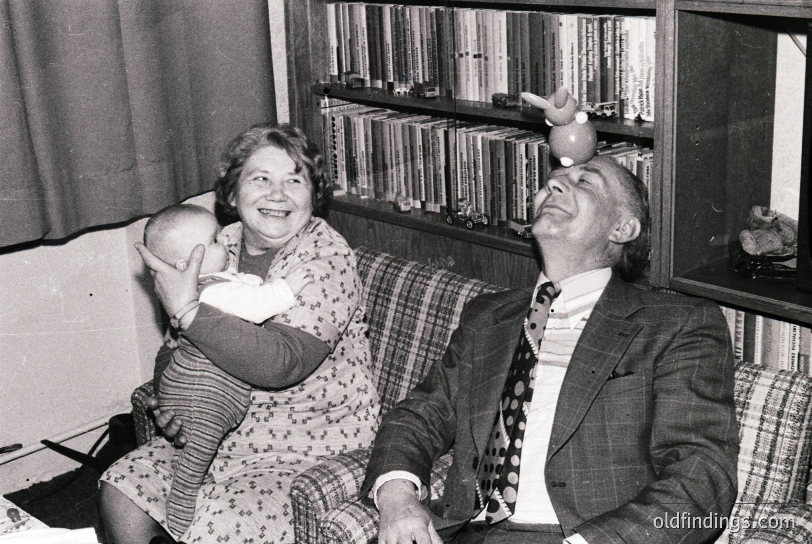 Mid-20th century black-and-white photo of two joyful adults in a cozy indoor setting. Woman in floral dress cradles a baby, man in suit lifts a toy bird above his head. Bookshelves filled with volumes line the background, suggesting a scholarly or family home. Likely 1950s–1960s, Western or European setting.