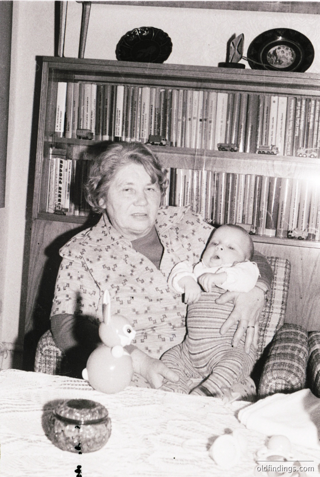 Mid-20th century indoor portrait of an elderly woman cradling a baby in a modestly furnished room. Wooden bookshelves filled with books and decorative items line the background. Floral-patterned blouse and striped baby garment suggest domestic warmth. Vintage