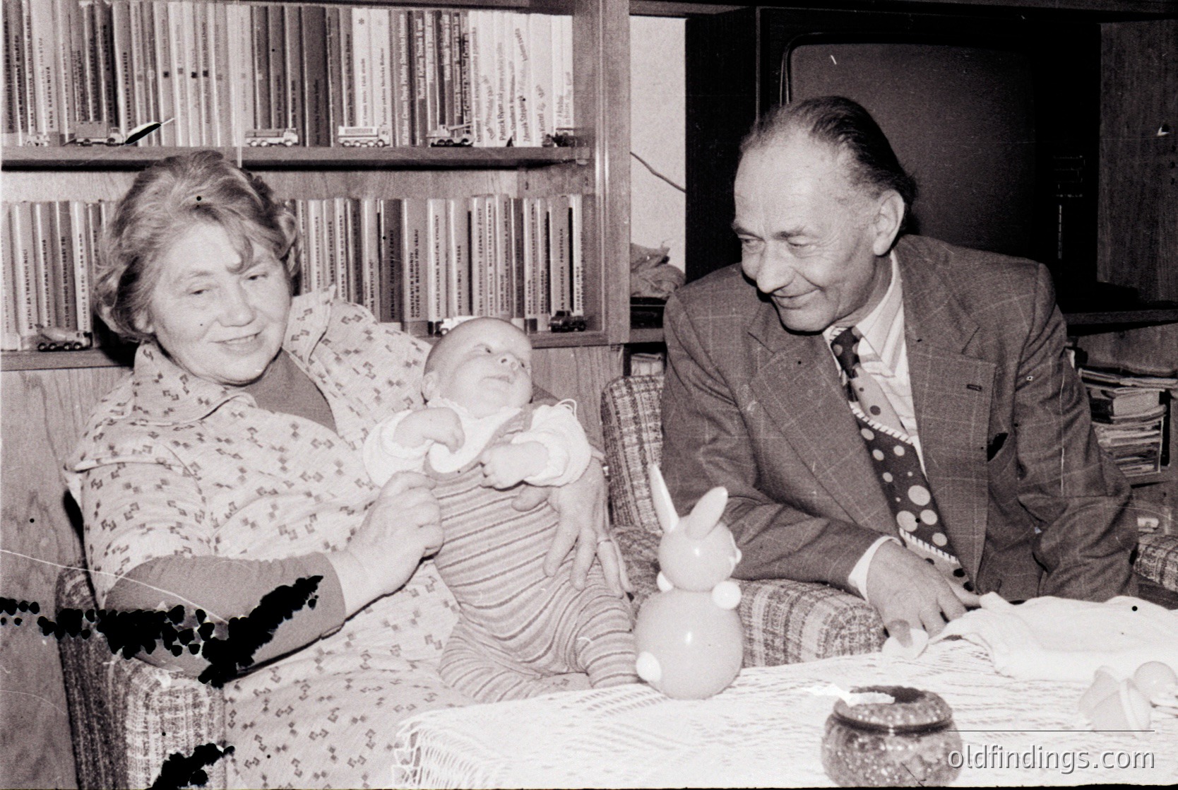 A mid-20th century (likely 1950s–1960s) indoor portrait showing an elderly couple with a baby. The woman, dressed in a floral blouse, holds a striped baby in a striped dress. The man, in a suit with a patterned tie, sits beside her. A bookshelf with bound volumes and a ceramic bunny figurative vase fill the background. Warm, intimate family moment.