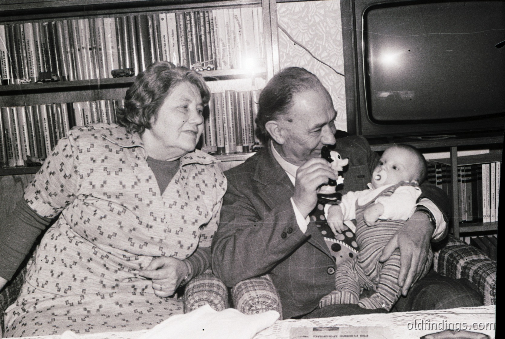 Black-and-white interior shot of elderly couple sharing a moment with a baby, likely mid-20th century. Man in suit holds baby while woman in floral dress leans in, suggesting familial warmth. Old-fashioned TV in background and vintage furniture indicate or domestic life. Ideal for historical research or nostalgic design references.
