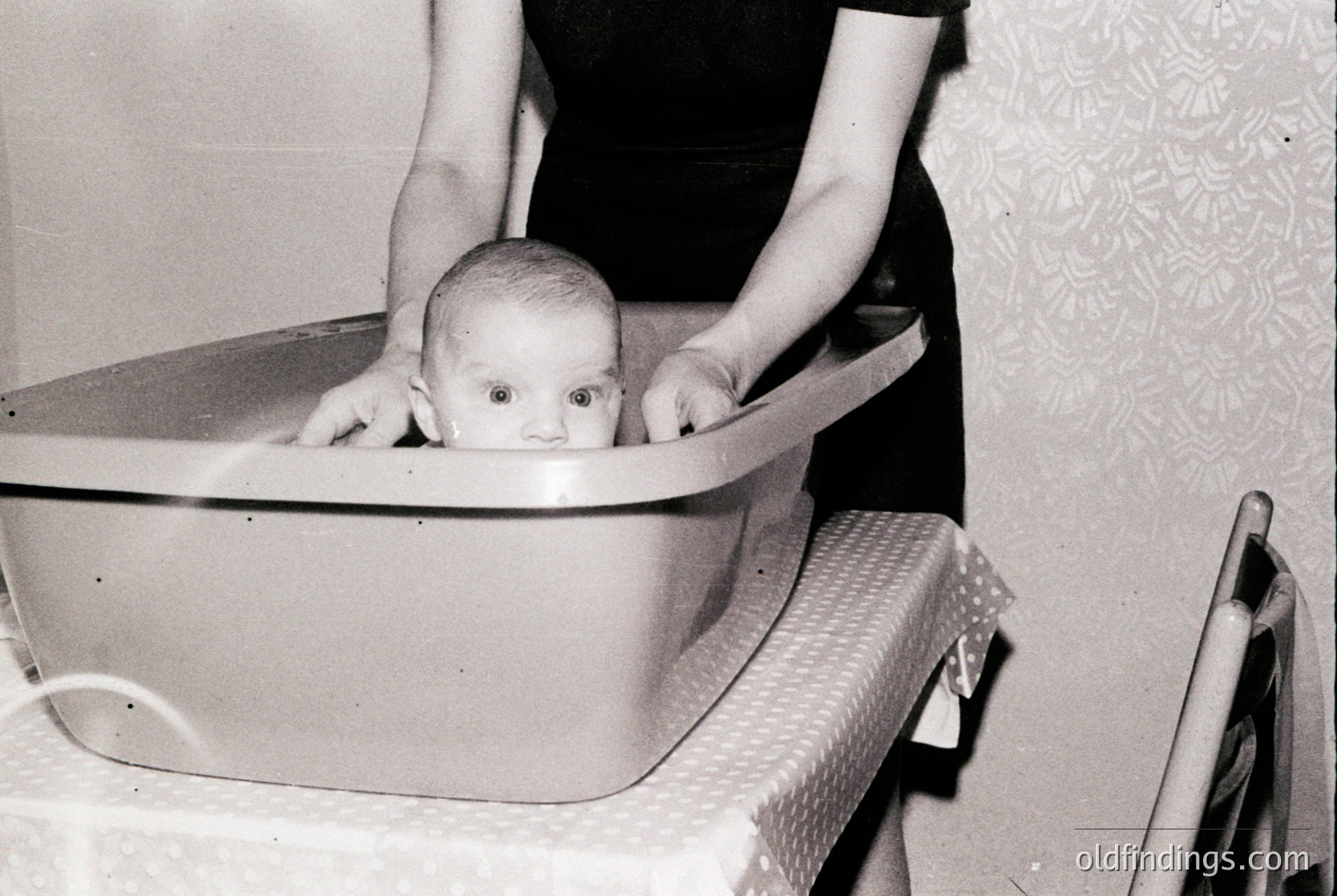 Mid-20th century indoor scene: infant in a metal baby tub, held by an unseen adult’s hands. Patterned wallpaper with floral motifs suggests domestic setting. Tub rests on a metal tray with wheels, likely a vintage bathing setup. Candid, nostalgic family moment.