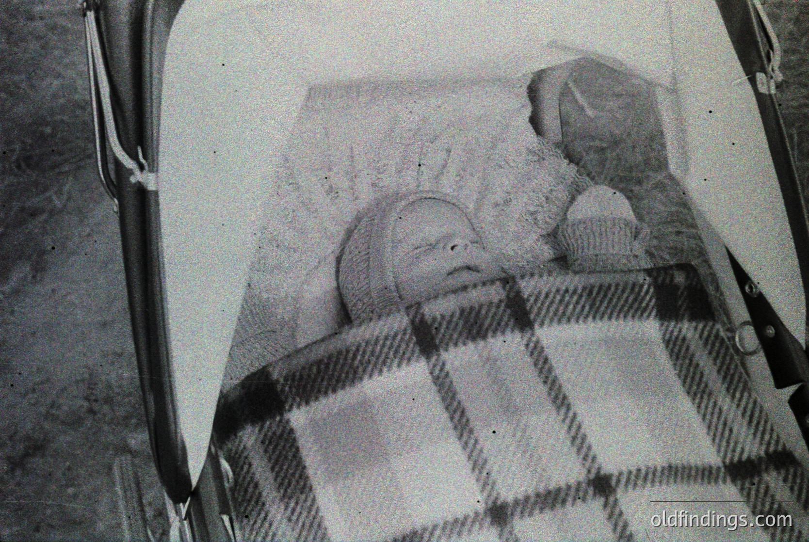 Vintage black-and-white photo of an infant sleeping in a vintage pram with checkered blanket and knitted cap. Cupcake beside the child suggests a celebratory or casual moment. Likely mid-20th century, possibly 1950s–1960s.