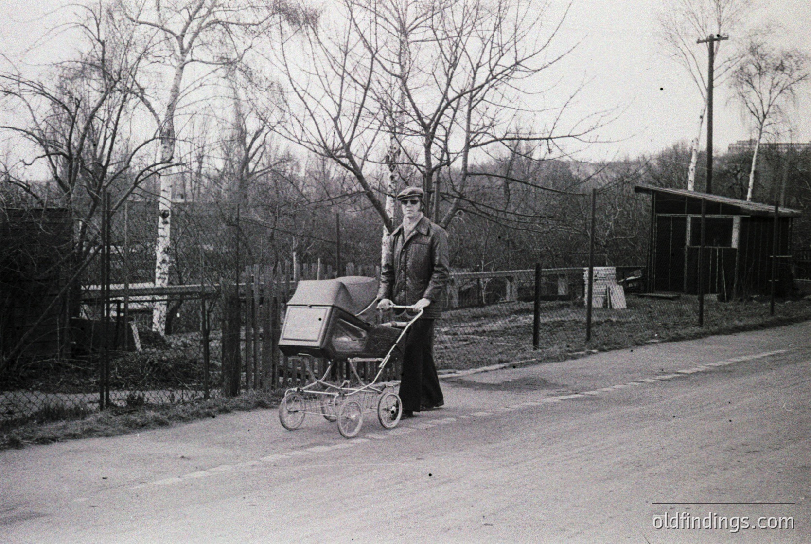 A woman in mid-20th century attire pushes a vintage pram along a quiet, tree-lined road. Her long coat and hat suggest a European urban/suburban setting, likely . The barren trees and paved road hint at winter or early spring. Ideal for historical research on family life or vintage fashion.