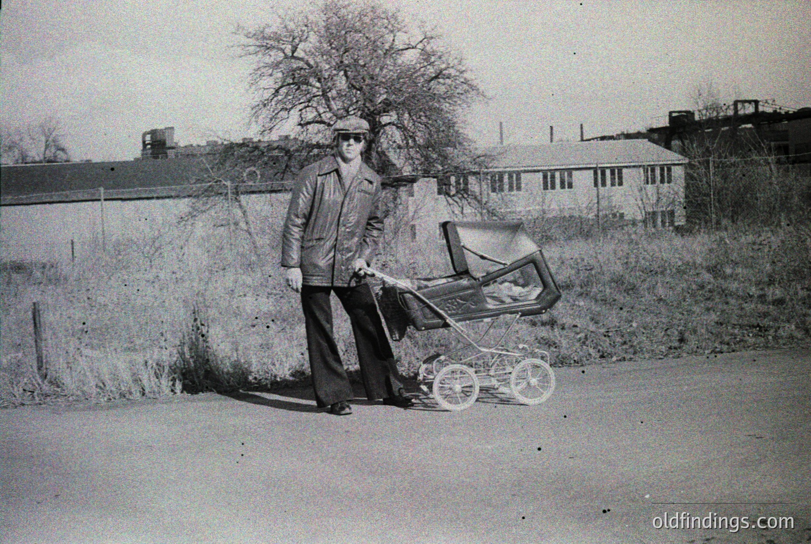 Man in 1970s-style wide-leg trousers and cap pushes a vintage pram on a rural road, surrounded by sparse trees and industrial buildings. Classic mid-century urban/suburban lifestyle captured.