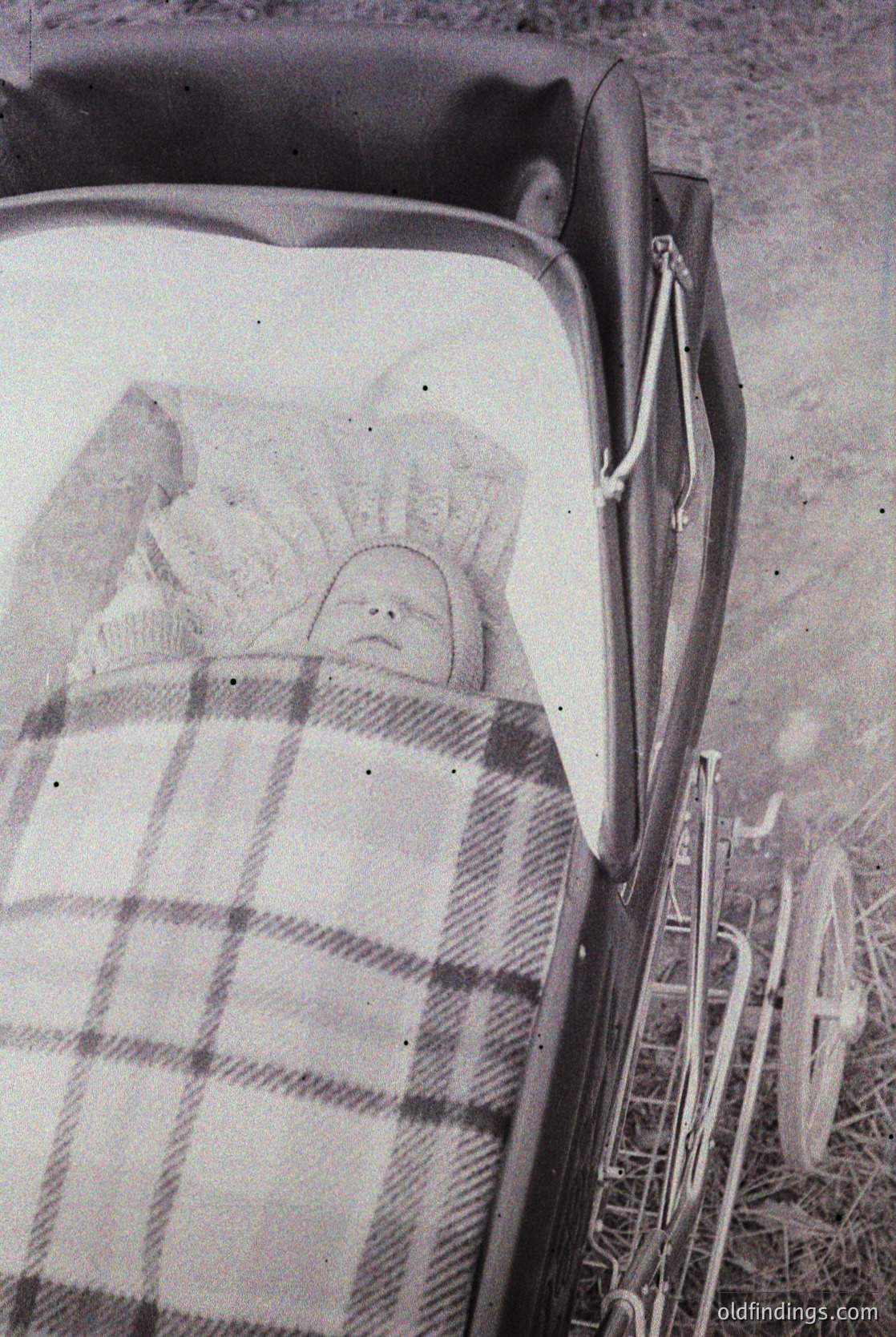 Vintage black-and-white photo of an infant wrapped in a plaid blanket inside a vintage stroller, likely mid-20th century. The stroller’s metal frame and curved handlebars suggest mid-century design. Outdoor setting with blurred greenery in background.