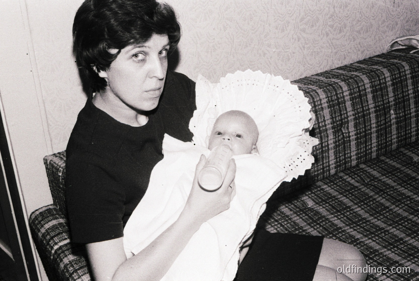 A woman in a short-sleeved blouse cradles an infant wrapped in a lace-trimmed bib, feeding them from a small glass bottle. The scene appears indoors, likely mid-20th century, with a plaid-patterned armchair and floral wallpaper. The composition captures a tender, candid moment of domestic life.