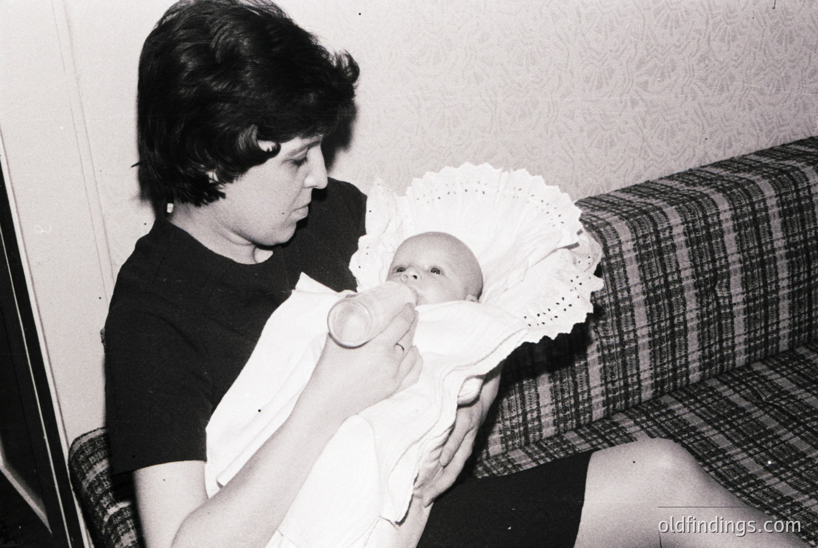 A woman feeds a swaddled infant in a mid-20th-century domestic setting. Floral wallpaper and a plaid couch suggest 1960s–1970s decor. The infant wears a lace-trimmed bib, common in mid-century parenting practices.