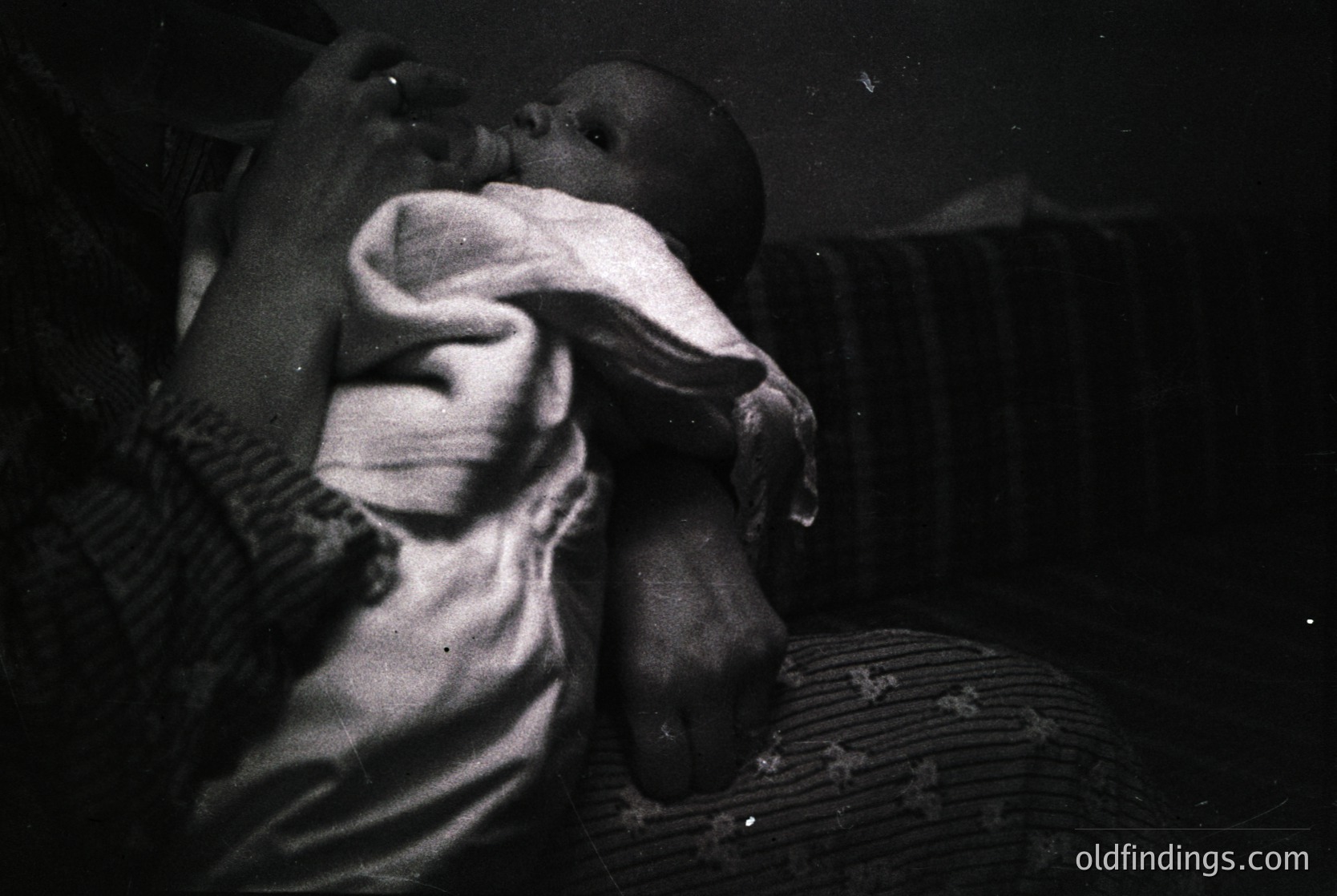 Black-and-white candid of an adult cradling an infant, likely mid-20th century. Soft focus and low lighting emphasize tender, intimate moment. Textured floral fabric and striped sleeve detail add warmth. Ideal for historical research on parenting or vintage photography.