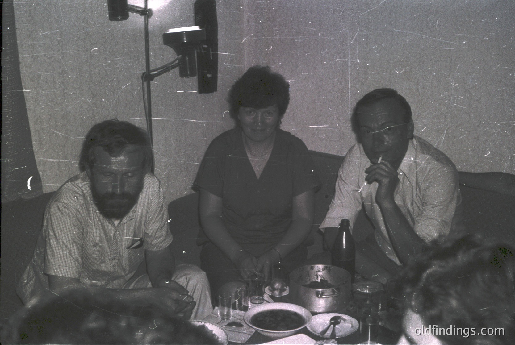 Three individuals seated at a table in a dimly lit studio setting, likely mid-1970s. The man on the left wears a striped button-down, the woman in the center a collared blouse, and the man on the right a light shirt with glasses. A camera mounted on a tripod suggests staged portraiture. Empty plates and cutlery hint at a meal setting.