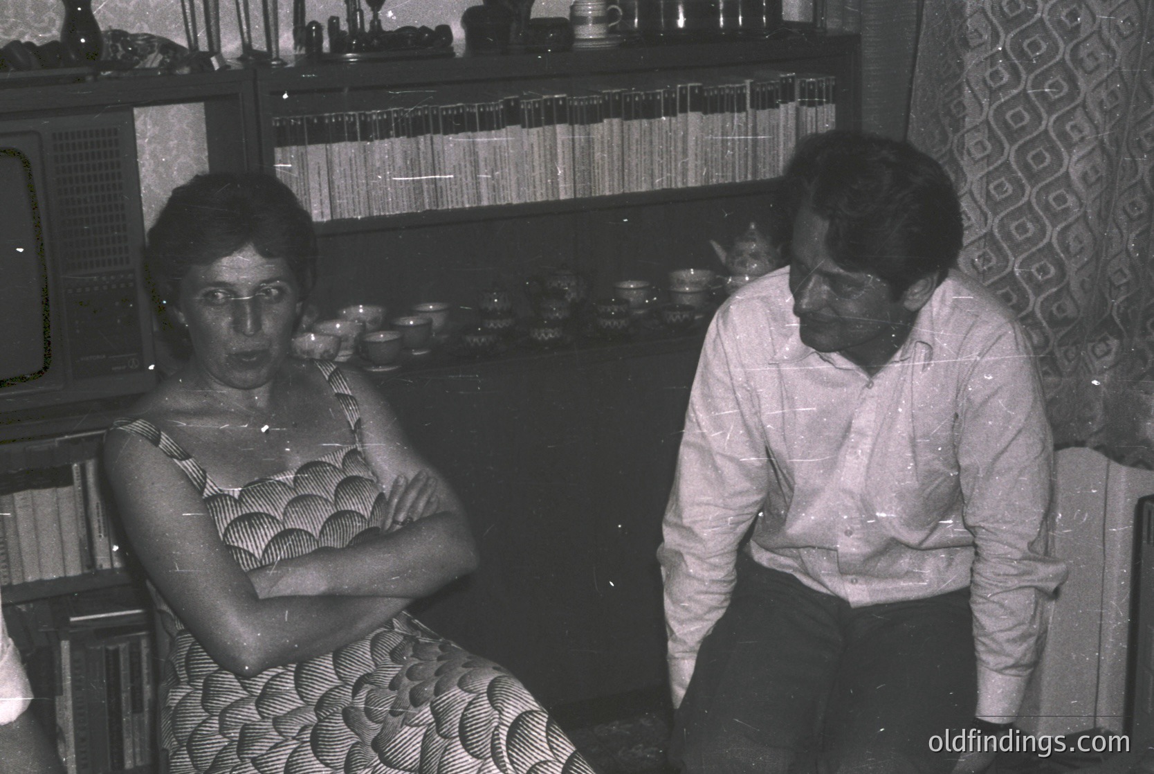 Two individuals seated indoors in a mid-20th-century setting, likely 1960s–1970s. Woman wears a patterned dress with geometric motifs; man in a button-down shirt. Shelves behind them display vinyl records, glassware, and decorative items. Warm, intimate domestic atmosphere.