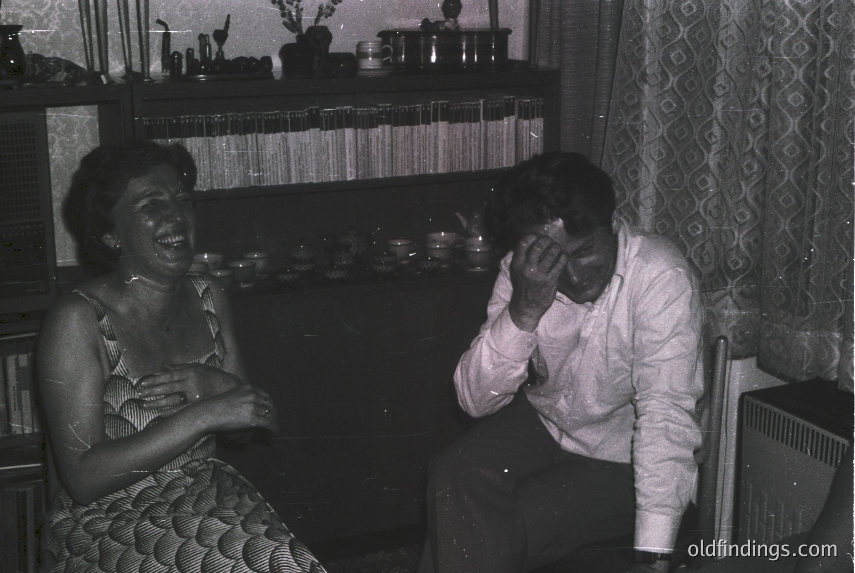 Vintage black-and-white indoor scene of two people laughing in a mid-century living room. Woman in a patterned dress sits left, man in a collared shirt reacts playfully right. Shelves behind display glassware and books; floral-patterned wallpaper adds warmth. Likely 1960s–1970s domestic setting.