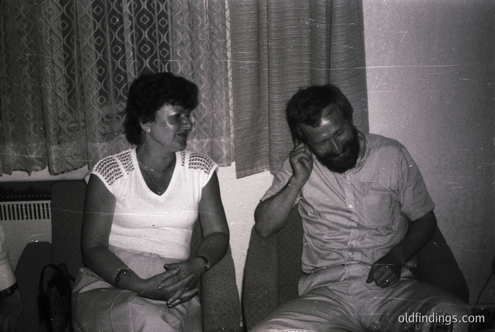 Two individuals seated indoors in a mid-20th-century setting, likely 1970s–1980s. Woman in a sleeveless blouse with geometric-patterned curtains behind; man in a striped shirt, resting chin on hand. Plain wall and radiator visible. Candid, intimate moment.