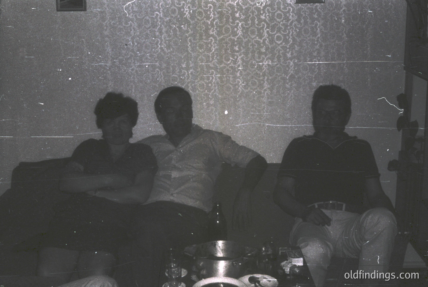 Three individuals pose indoors on a patterned sofa, likely mid-20th century (1960s–1970s). The woman wears a sleeveless dress, the man in the center a short-sleeve shirt, and the man on the right a collared shirt with rolled sleeves. A coffee table with a metallic tray sits between them. Decorative wallpaper features geometric motifs.