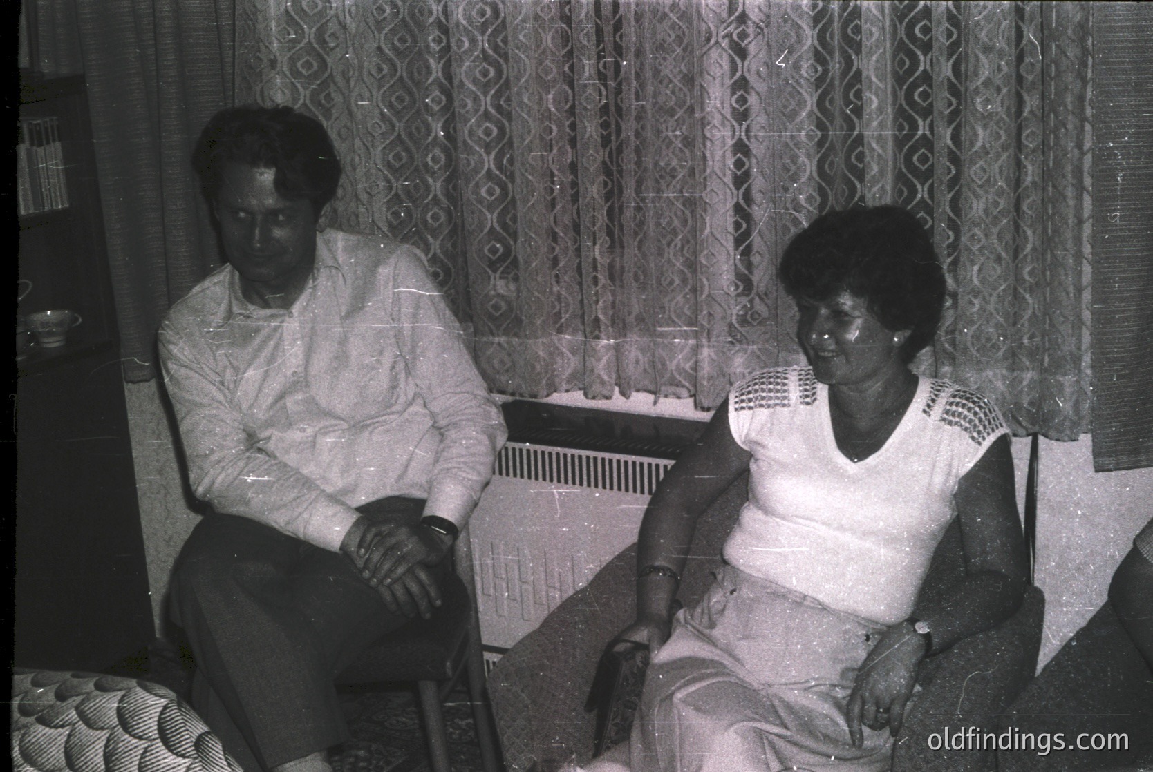Two individuals seated indoors in mid-20th century attire, likely 1960s–1970s. Man in button-down shirt, woman in patterned blouse with high-neck collar. Patterned curtains and vintage radiator visible. Warm, intimate domestic setting.