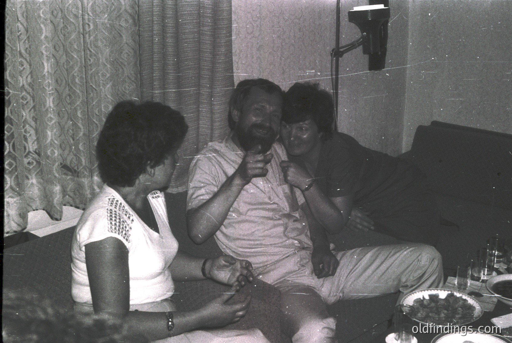 Vintage indoor gathering featuring three adults in casual 1970s attire—man in a light-colored button-up shirt, woman on left in a white blouse with lace trim, woman on right in a dark top. Setting includes a tiled wall, patterned curtains, and a table with food/drinks. Candid moment captures relaxed social interaction.