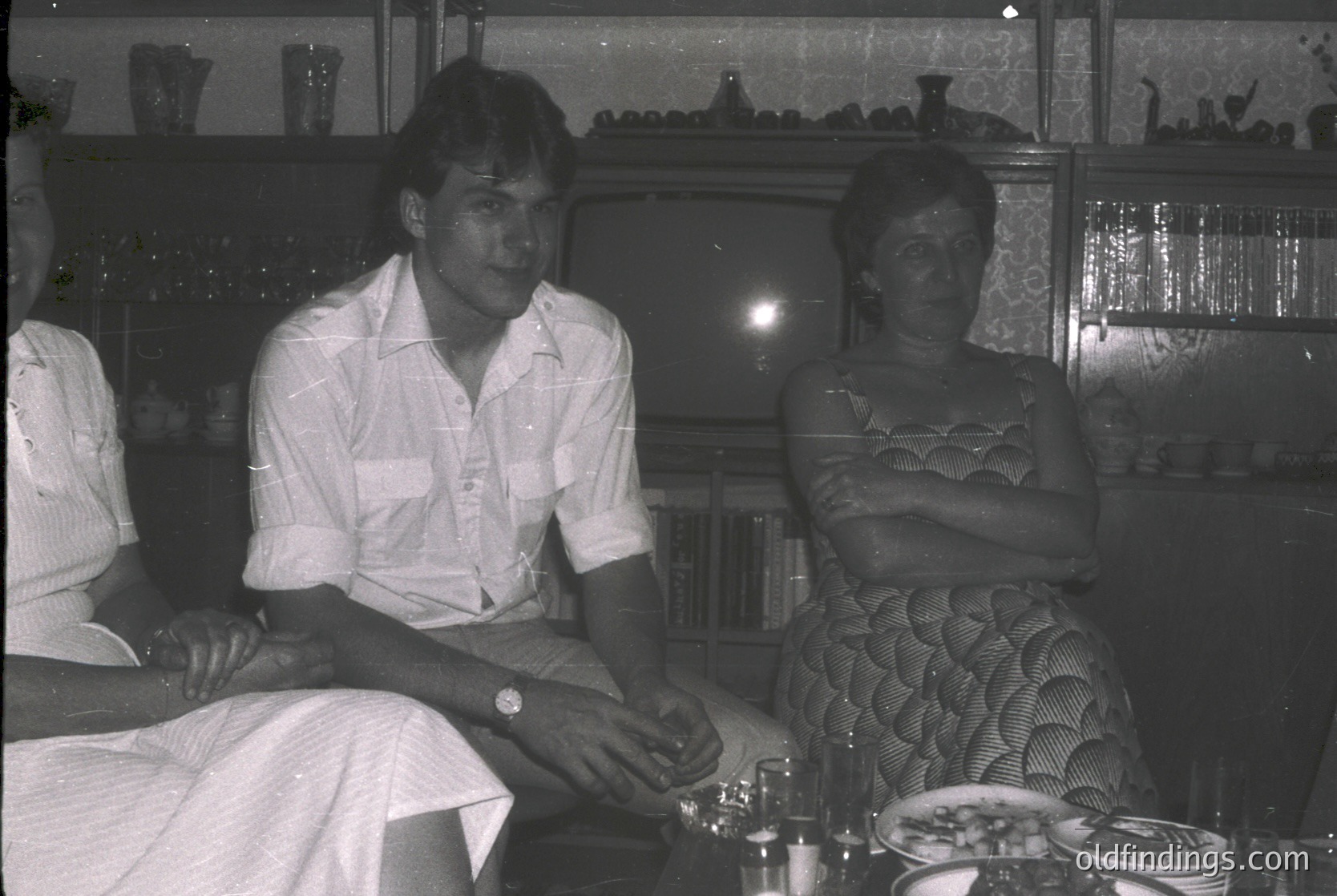 Black-and-white interior shot of three individuals seated at a table in a dimly lit room, likely a 1970s café or restaurant. The man wears a short-sleeve button-down shirt, while the woman’s patterned dress features a geometric, puckered design. Glassware, plates, and decorative shelves with glassware in the background suggest a mid-century social setting. éCulture