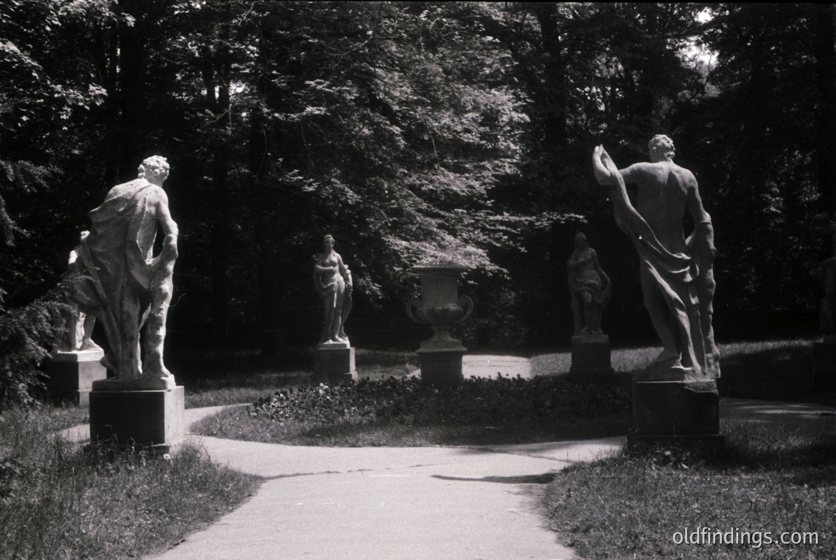 Neoclassical stone statues flanking a sunlit pathway in a lush garden, likely 19th–early 20th century. Four figures—two male (one gesturing, one standing), two female—positioned on rectangular pedestals. Dense foliage and a central plaque suggest a formal park or estate setting. Ideal for historical research, architectural studies, and vintage design references.