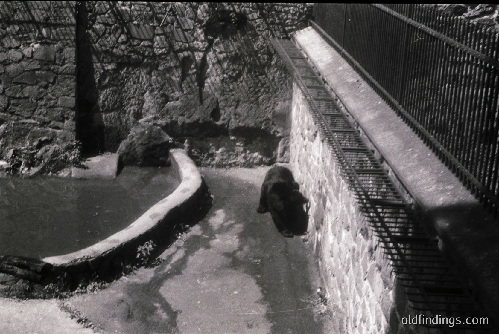 Industrial-era water reservoir with curved concrete basin and metal grating. A lone worker in dark attire stands near a sloped drain, surrounded by stone and brick walls. Likely a mid-20th century factory or utility site.