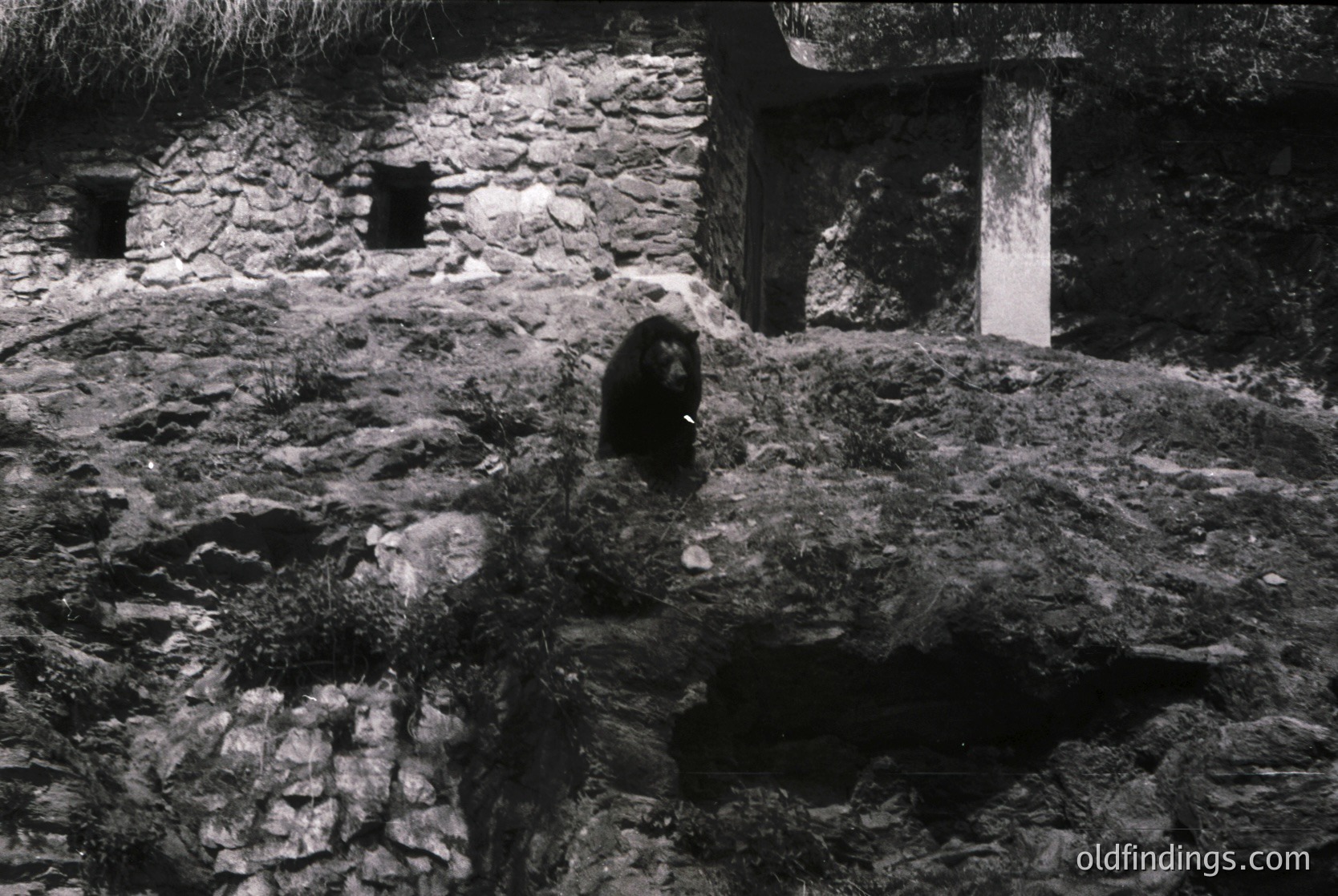 Black-and-white photograph of a person seated on moss-covered stone steps in a dimly lit, rustic courtyard. Stone walls and arched openings frame the scene, suggesting historic architecture. The individual wears a dark, loose-fitting garment, possibly traditional or utilitarian.