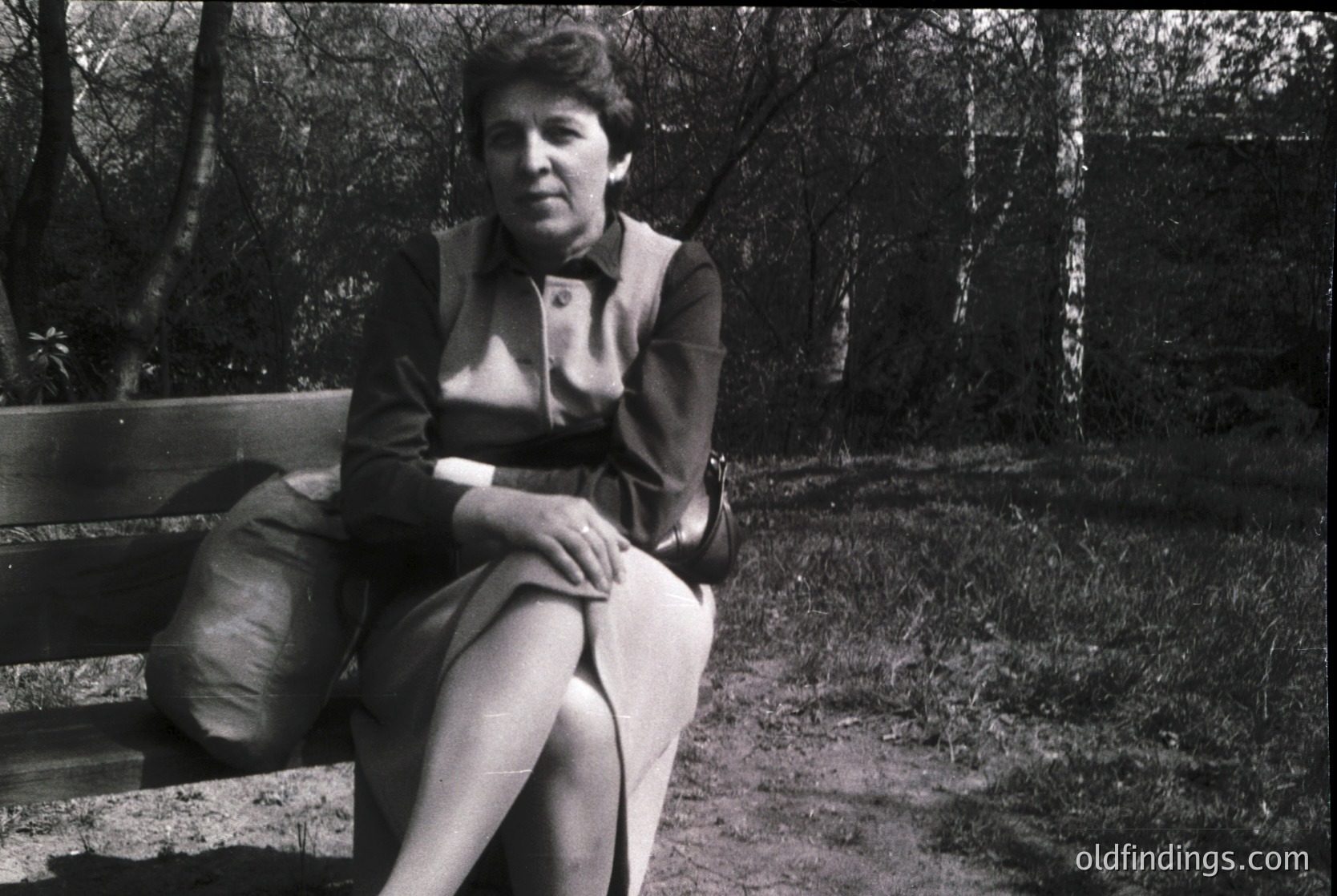 Black-and-white portrait of a woman seated on a wooden bench in a forested area, mid-20th century. She wears a structured blazer, striped blouse, and trousers, suggesting a formal or professional setting. The dense foliage and dirt path indicate an outdoor, possibly rural or park-like environment.