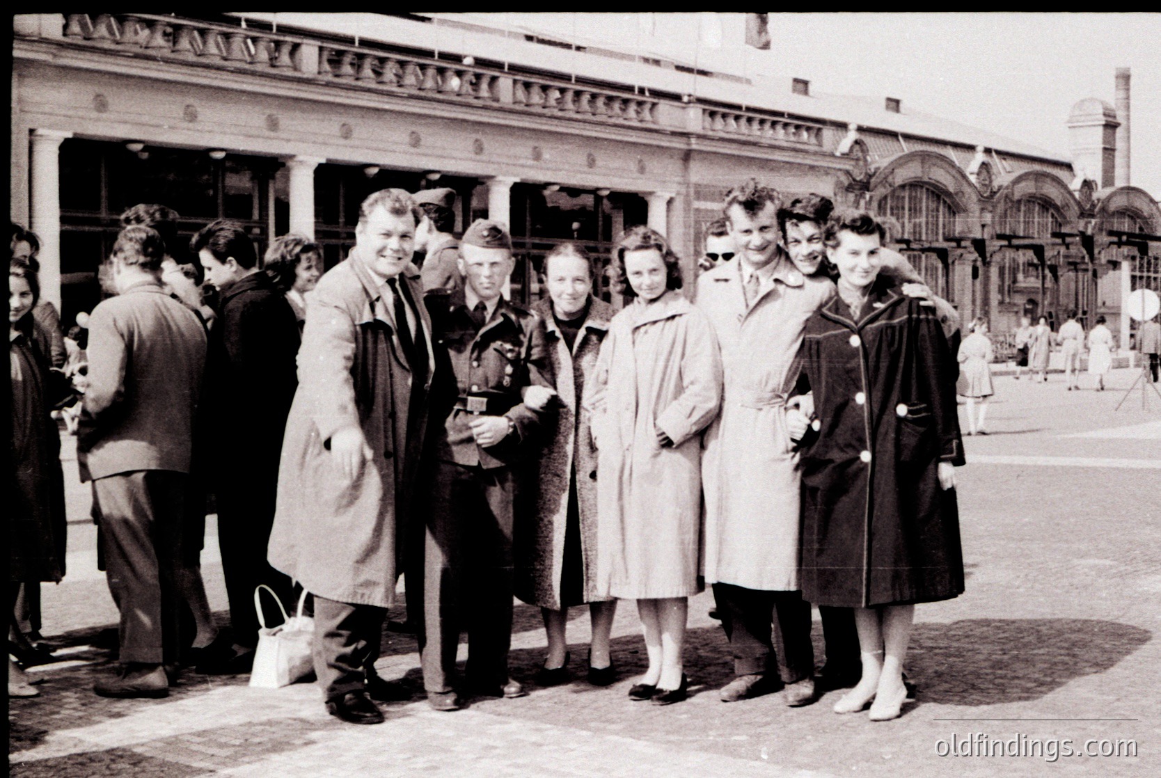 Mid-20th century group photo outside grand neoclassical train station. Seven individuals in 1950s attire—men in suits/coats, women in knee-length coats & gloves—pose formally. Architectural details include arched windows, columns, and decorative cornices. Crowd of onlookers in background.
