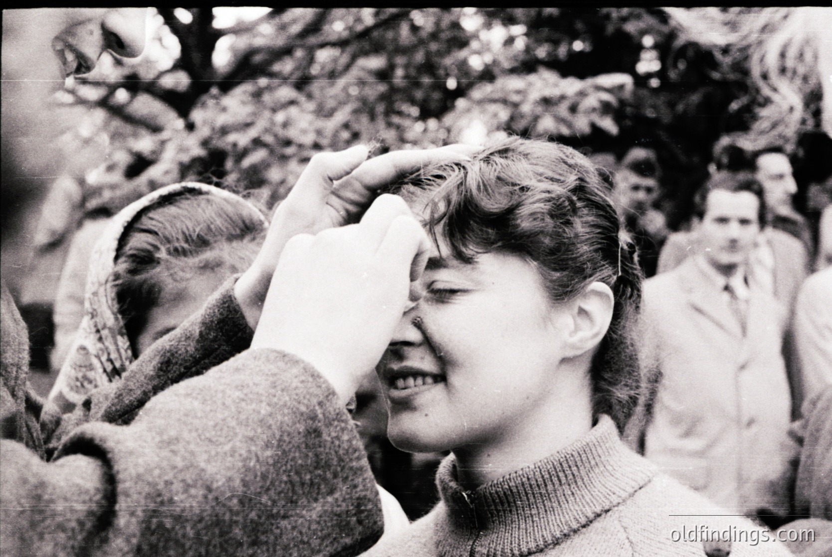 Mid-century street scene: A woman in a turtleneck sweater adjusts her hat in a crowd, surrounded by blurred figures in winter coats. Likely 1950s–1960s urban setting. Candid, documentary-style composition.