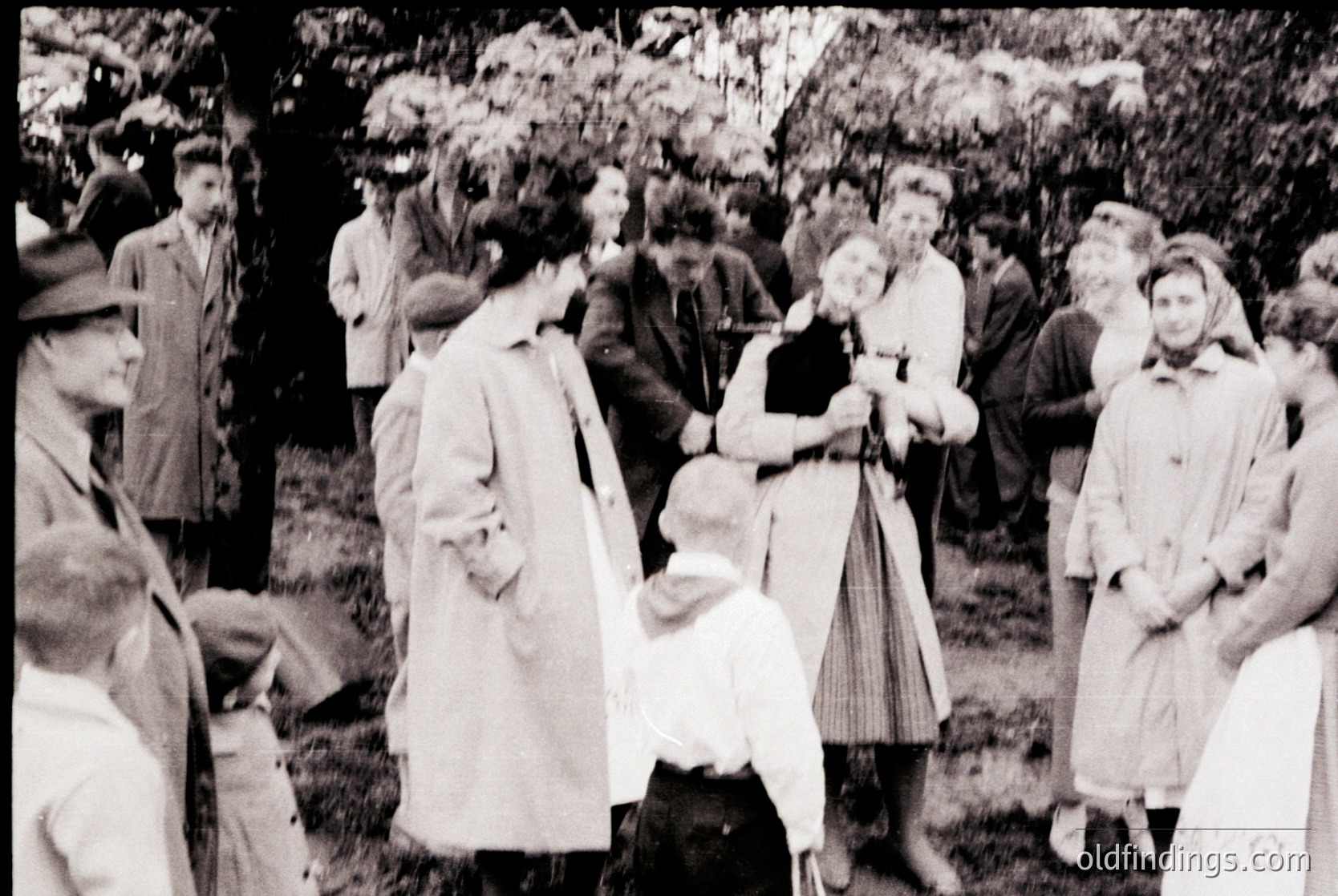 Vintage black-and-white outdoor gathering, likely mid-20th century. Crowd in autumn foliage, featuring men in suits, women in long dresses and coats, and a child in a light dress. Central figures appear to be embracing or greeting. Rural or park setting with trees and grass.