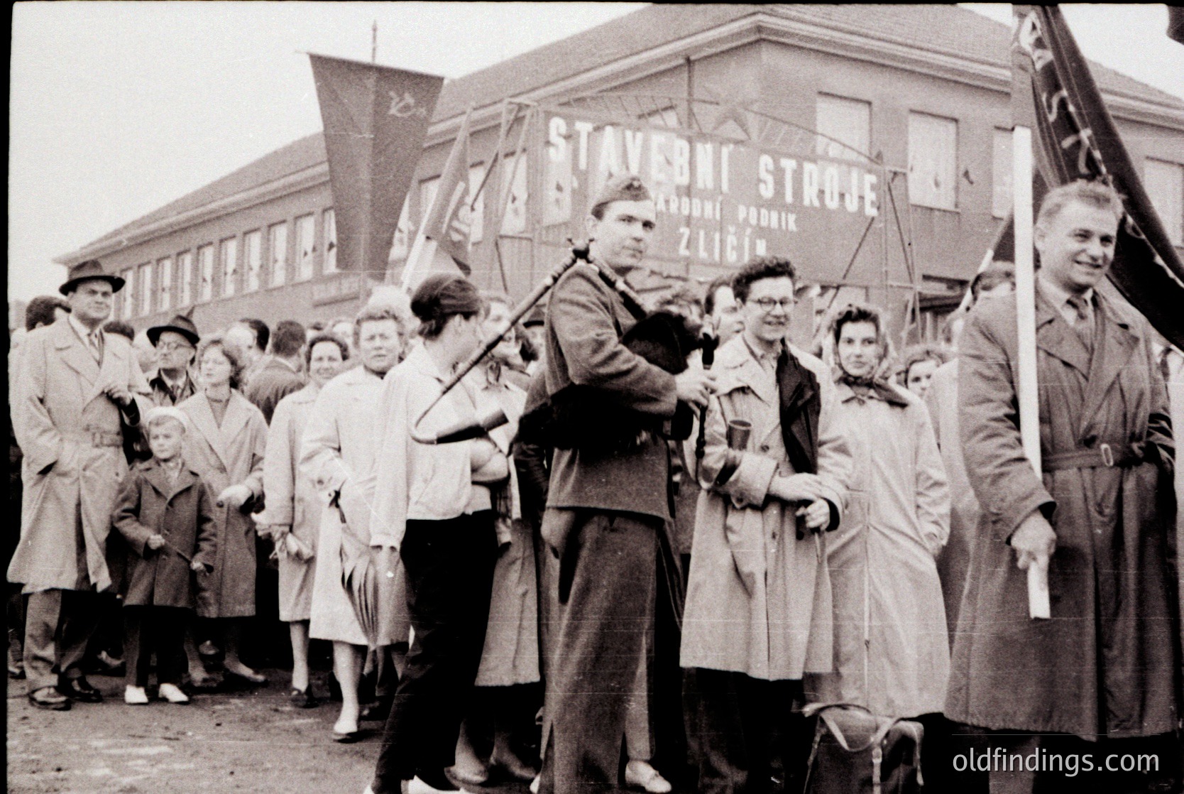 Black-and-white street scene featuring a crowd gathered outside a building with signage in Czech: "Stavební Společnost" (Construction Society) and "Zlín." Central figures pose with a hammer-and-sickle flag and a banner, likely from the 1940s–1950s. Men in military-style coats and women in long coats and hats dominate the crowd.