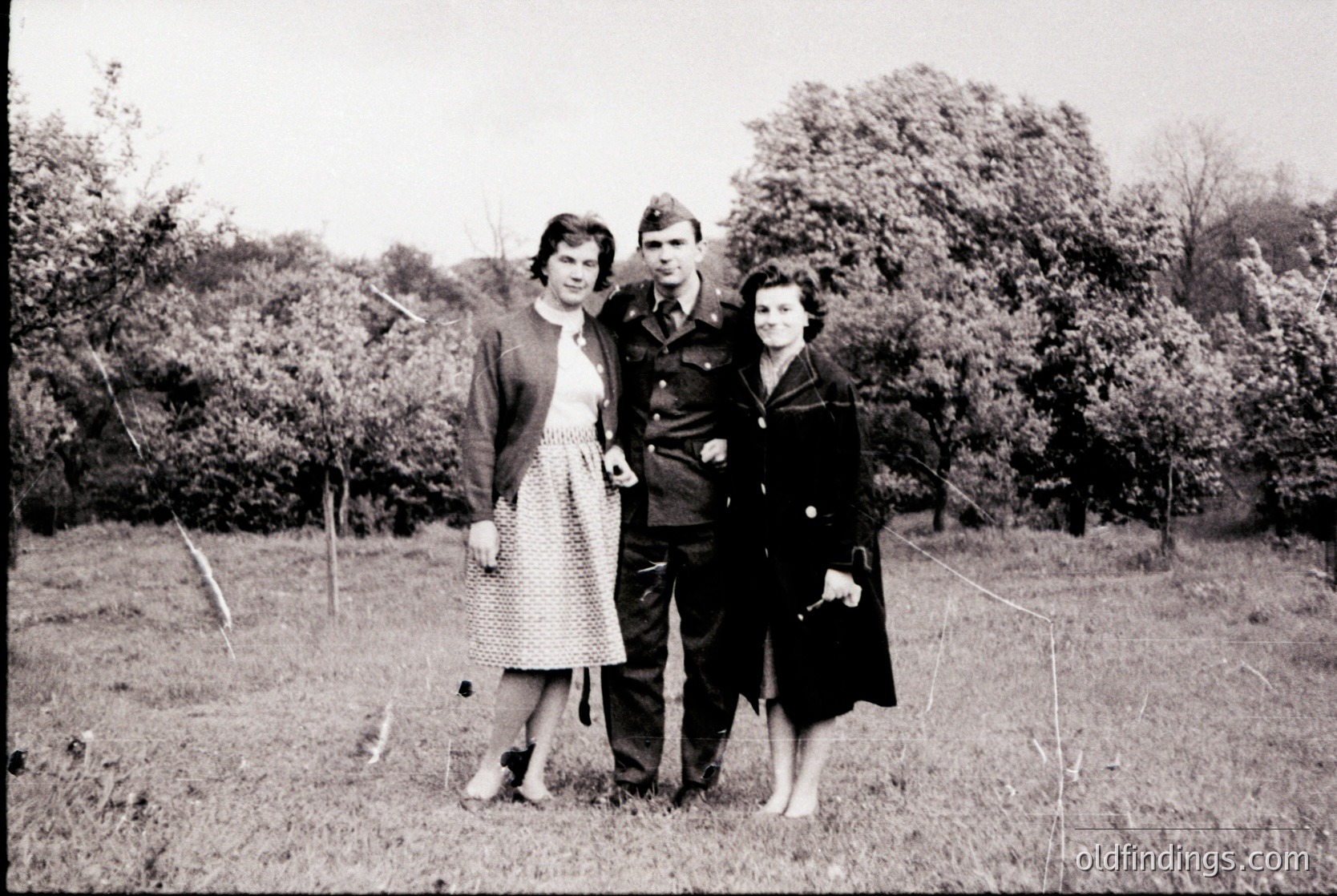 Three individuals pose outdoors in mid-20th-century attire, likely 1940s–1950s. The man wears a military uniform with insignia, flanked by women in patterned dresses and structured coats. Lush greenery and overcast skies frame the scene, suggesting a rural or park setting.