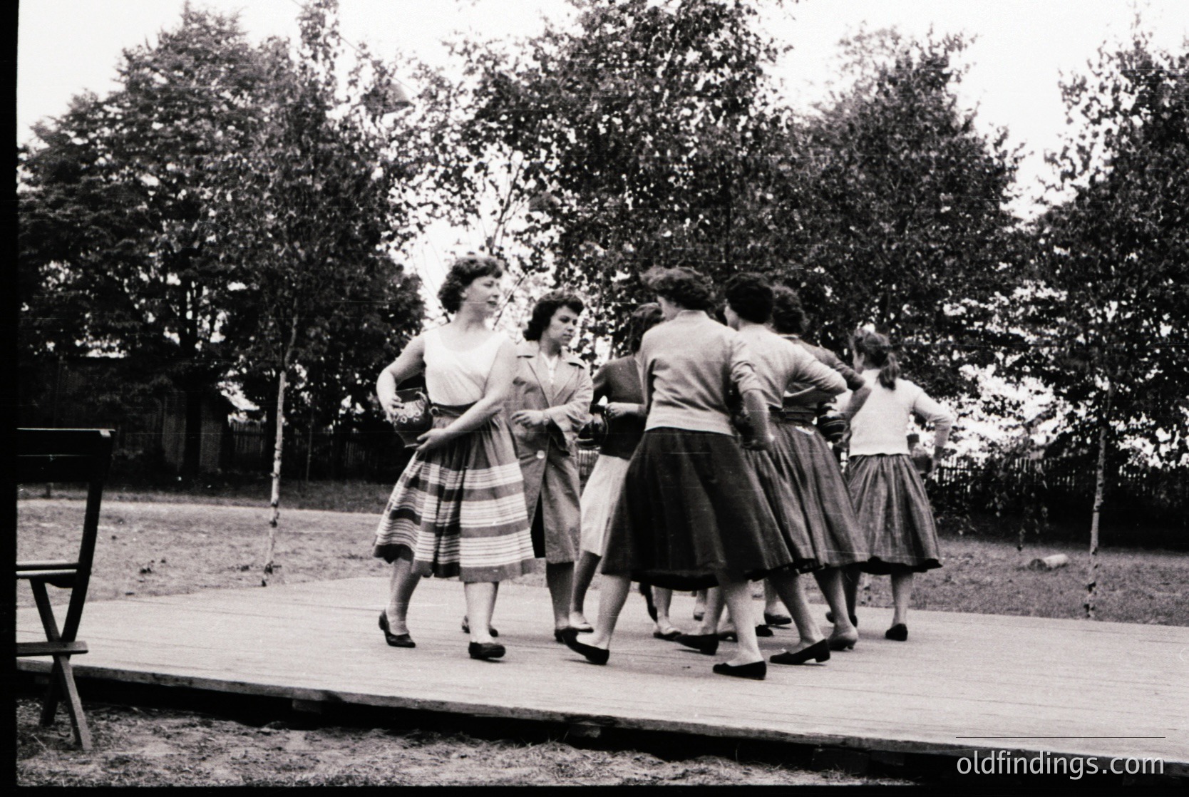 Group of women in 1950s-style dresses and pleated skirts performing outdoors on a wooden platform, likely a folk or dance ensemble. Mid-century fashion with fitted tops and full skirts. Wooden fence and trees in background suggest a rural or park setting.