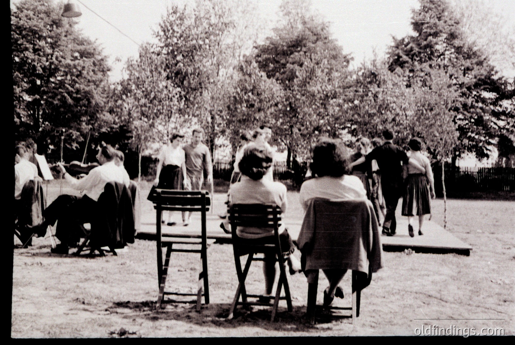 Mid-20th century outdoor gathering in a park setting, featuring women in long skirts and headscarves playing croquet. Wooden chairs and a croquet mallet are visible. Trees and a fence frame the scene, suggesting a communal or recreational event. Likely Eastern European or Soviet-era due to attire and setting.