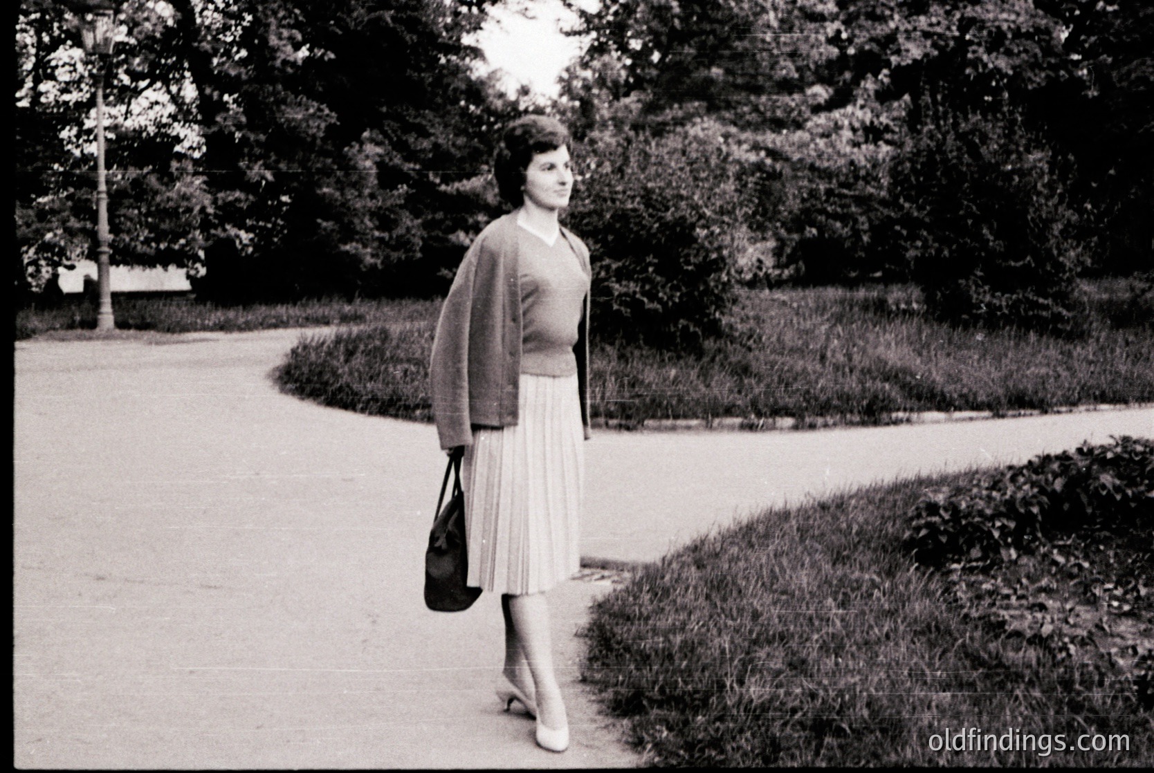 Mid-century woman in a fitted cardigan and pleated skirt strolls along a tree-lined park path, holding a structured handbag. Soft focus and high-contrast black-and-white style suggests 1950s–1960s fashion and photography. Urban park setting with manicured greenery and paved walkway.