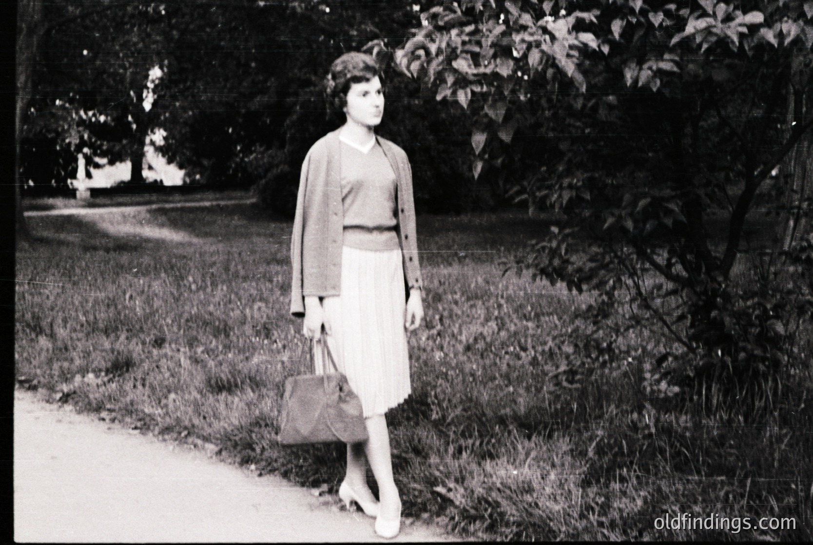Mid-century woman in a knee-length skirt and cardigan strolls along a tree-lined park path, holding a structured handbag. Soft focus and vintage monochrome suggest 1950s–1960s Western Europe or North America. Ideal for fashion history, retro design, or nostalgic stock imagery.
