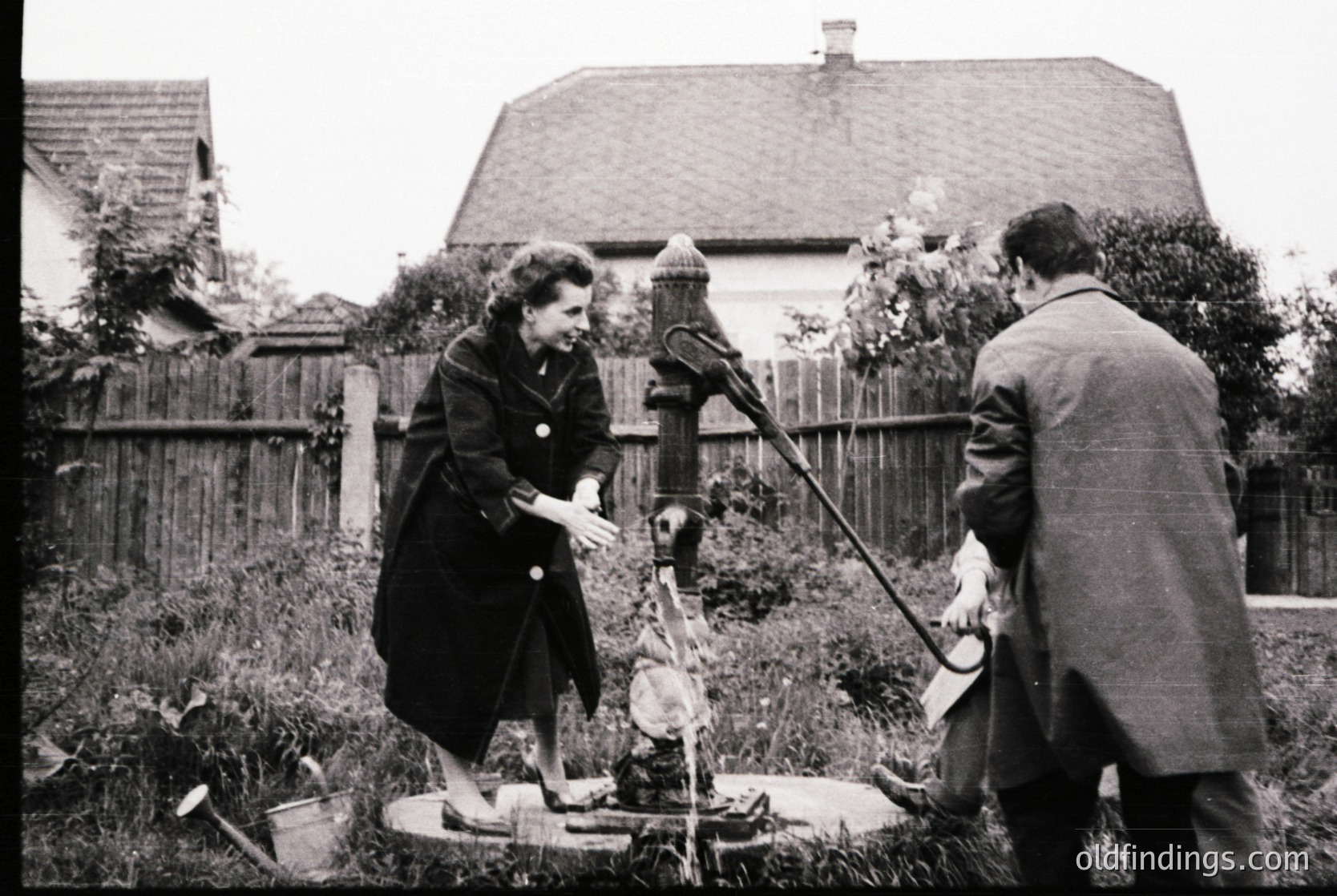 Mid-20th century (likely 1950s–1960s) black-and-white photo showing two individuals operating a manual well pump in a residential backyard. Woman in dark coat and hat assists man in suit with the pump handle, while a bucket sits nearby. Wooden fence and house with gabled roof in background. Rural or suburban setting.