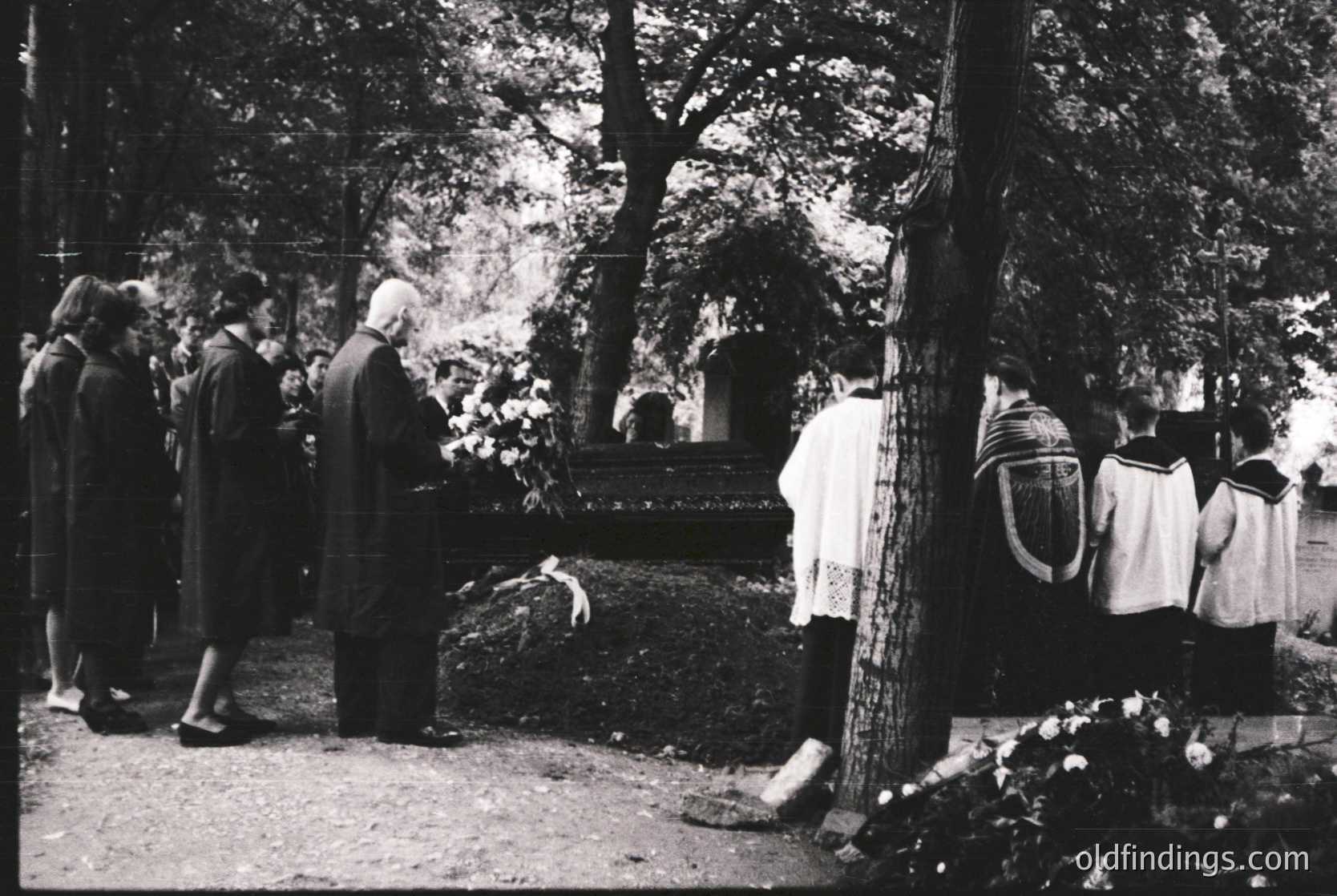 Black-and-white memorial scene in a forested cemetery, mid-20th century. Group of mourners, including a man in suit and women in traditional attire, gather around a grave adorned with floral tributes. Wooden cross and stone markers visible.