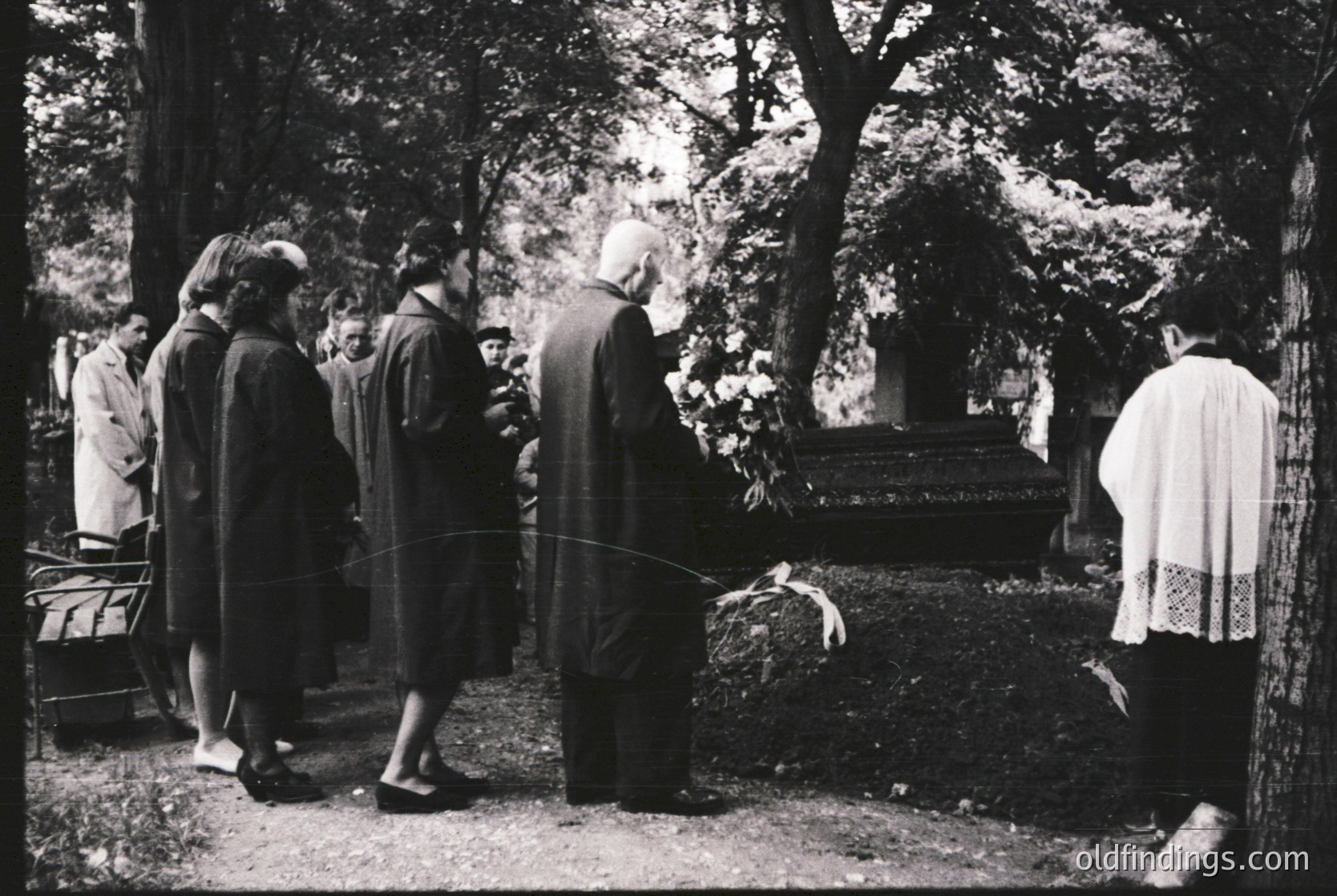 Mid-20th century funeral procession in a wooded cemetery. Group of mourners in formal attire—men in suits, women in long dresses—standing around a grave adorned with floral wreaths. Clergy in vestments conducts rites. Overcast forest backdrop with fallen leaves.