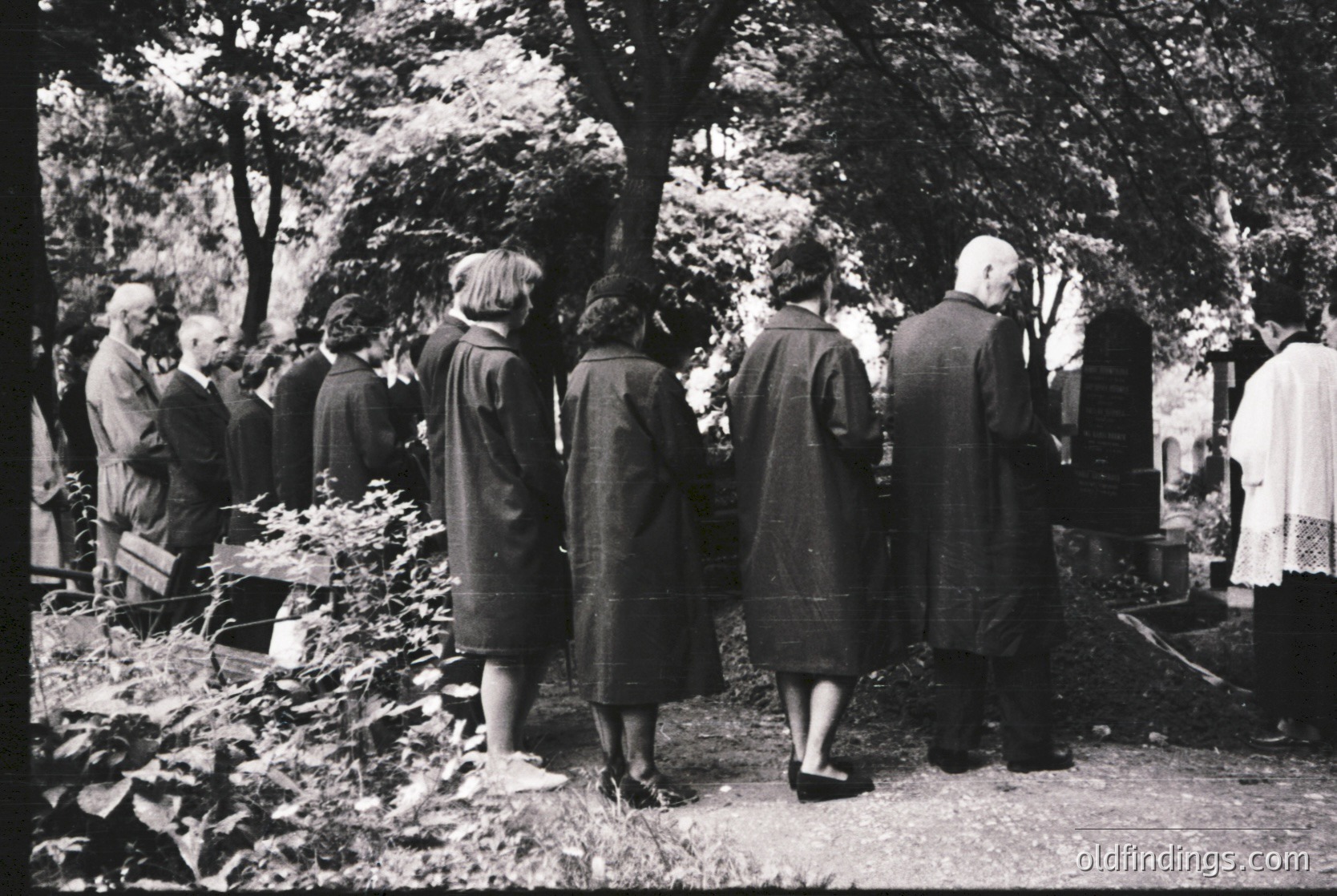 Black-and-white group standing at graveside in wooded cemetery, mid-20th century. Men in suits, women in long coats and hats, facing headstones. Overgrown foliage and stone pathway suggest rural or suburban burial ground. Formal attire indicates funeral or memorial service.