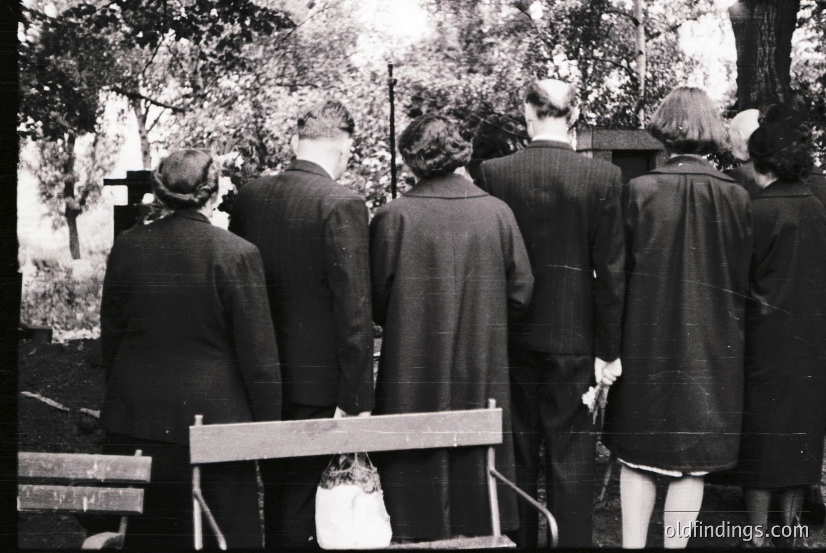 Group of six individuals in mid-20th century attire stands at a gravesite, facing crosses. Men wear suits, women in dresses with hats. Wooden bench and dog in foreground. Likely European cemetery, 1950s-1960s.