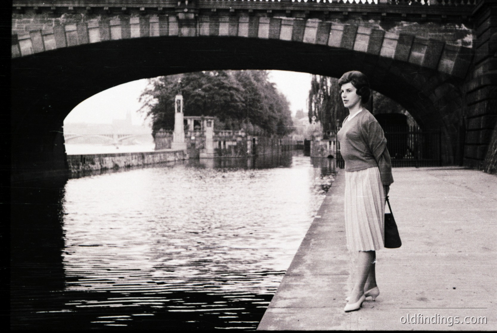 A woman in mid-20th-century fashion stands under a stone bridge arch beside a canal, holding a small handbag. Her skirt, cardigan, and hairstyle suggest the 1950s–1960s. The bridge’s brickwork and surrounding greenery hint at European urban design.