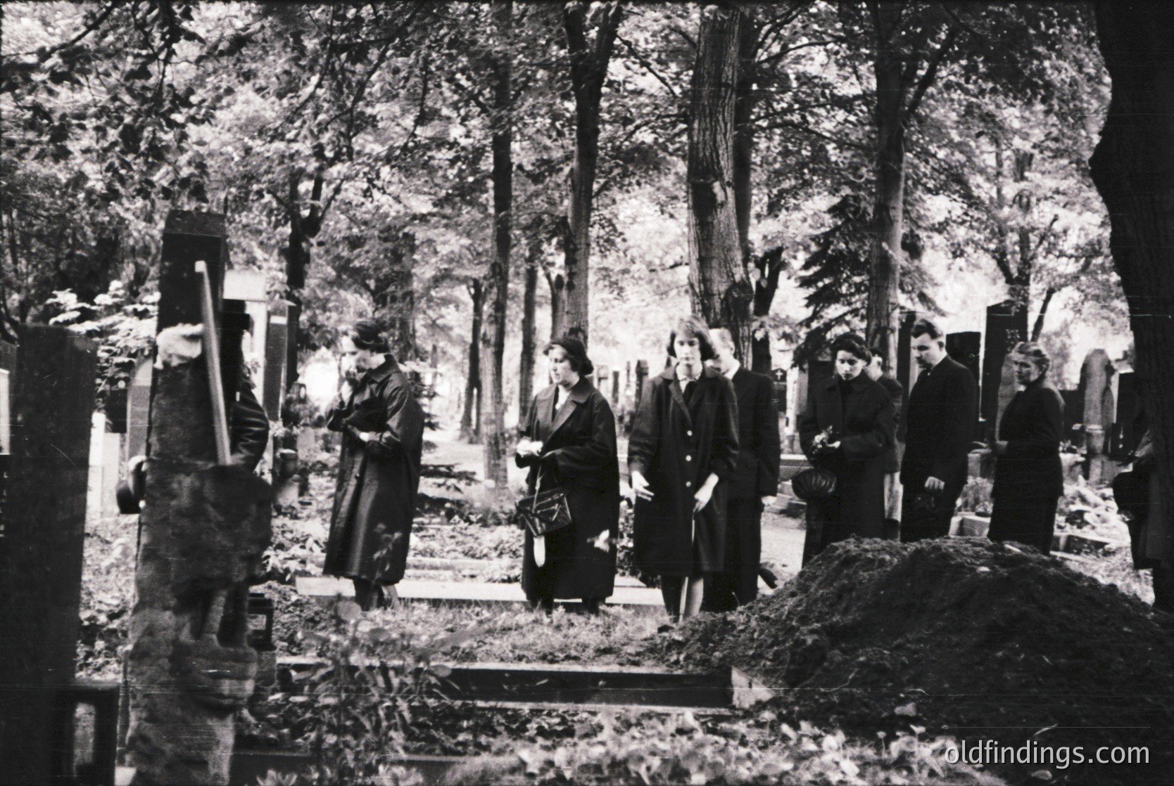 Black-and-white cemetery scene with seven mourners in traditional Eastern European attire (long coats, headscarves) gathered around a grave. Dense forest backdrop and weathered gravestones suggest rural setting, likely 19th–early 20th century.