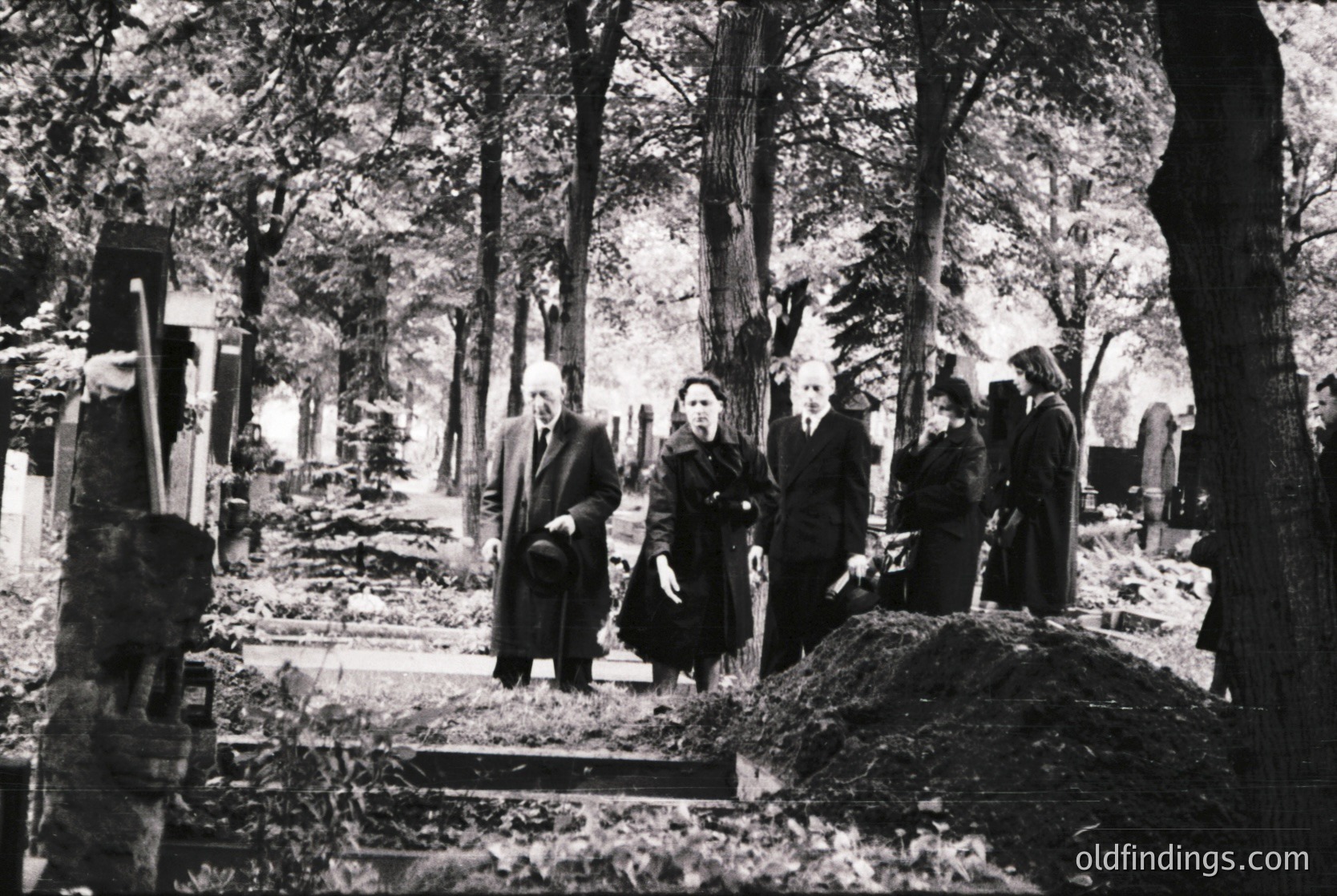 Mid-20th century black-and-white cemetery scene with overgrown graves and dense forest backdrop. Four mourners in dark clothing stand near a damaged headstone, suggesting wartime or post-war devastation. Clothing and setting evoke Eastern European or era.